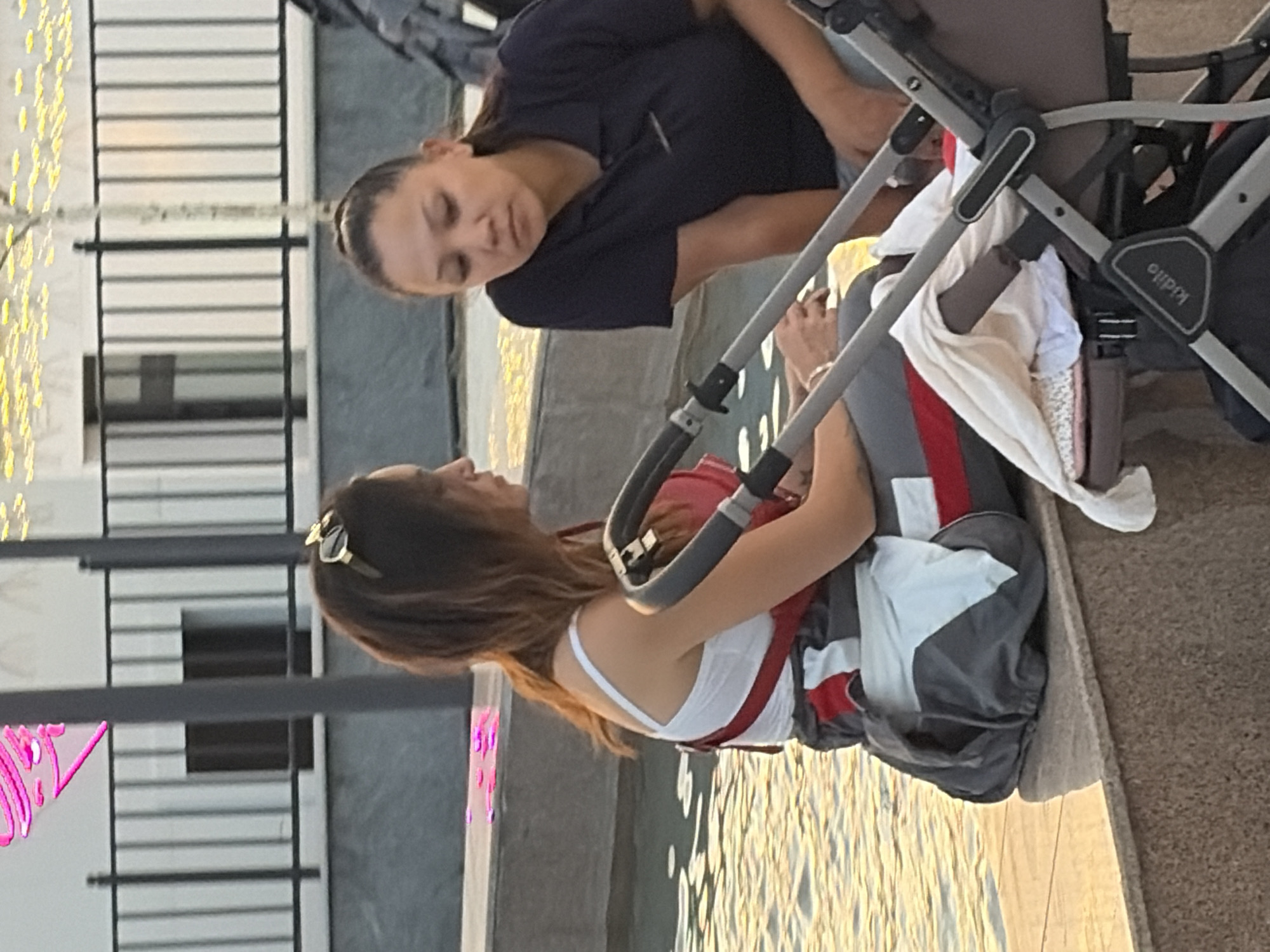 Two women are sitting on a ledge next to a body of water. The woman on the left is wearing a white tank top and grey overalls with red and white accents. She has brown hair and is looking to her right. The woman on the right is wearing a dark blue polo shirt and is looking down. They are both sitting near a stroller. The environment appears to be outdoors in a city setting, with a fence and lights visible in the background.