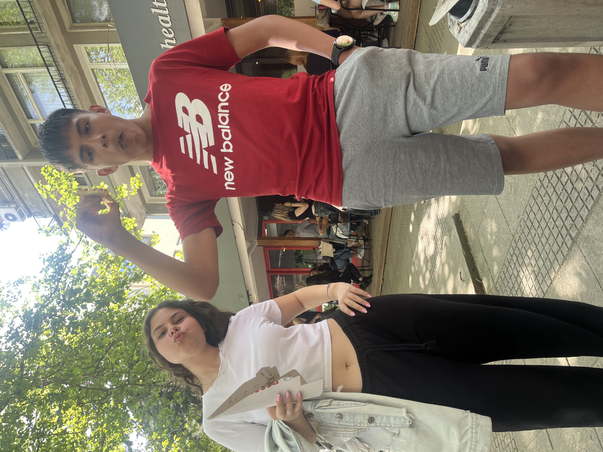 Two young people are posing for a photo on a city street, likely in Plovdiv, Bulgaria, given the location context. The male is wearing a red New Balance t-shirt and grey Puma shorts. He is holding something in his hand and making a face for the camera. The female is wearing a white t-shirt, black leggings, and holding a white, possibly cardboard object, along with a light denim jacket. She is also making a face for the camera. In the background, there is a building with a health-related sign and a restaurant with people seated inside and outside. The pavement is patterned. The sky is partly visible, with some trees adding greenery.