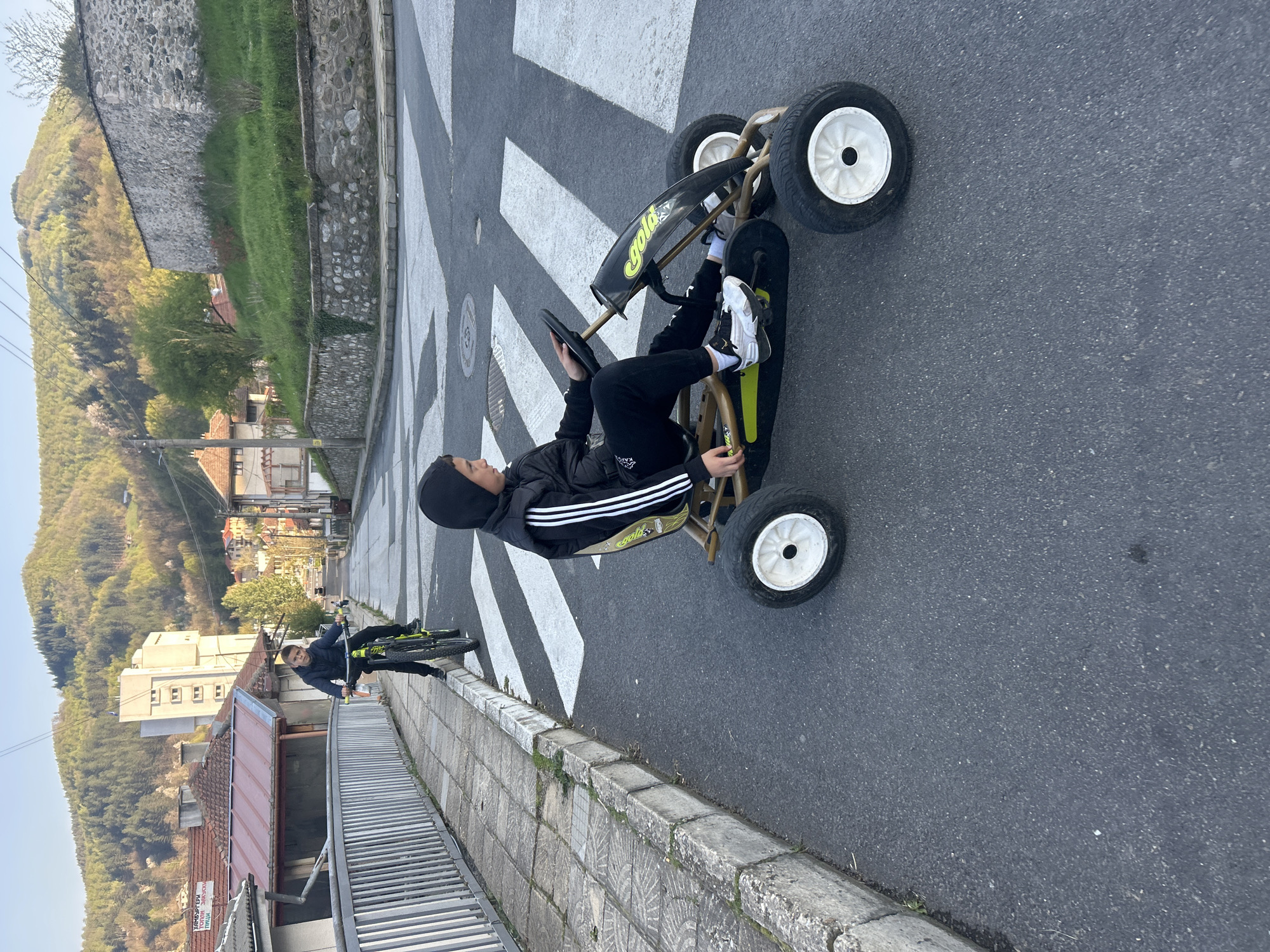 A young person wearing a black Adidas tracksuit and a black head covering is sitting in a "Gold" brand pedal go-kart on an asphalt road in Zlatograd, Bulgaria. The road has a crosswalk with white stripes. Another person is riding a bicycle behind them. The background features buildings, vegetation, and a hillside. The scene is captured during daylight. The buildings and vegetation suggest a residential area within the city.