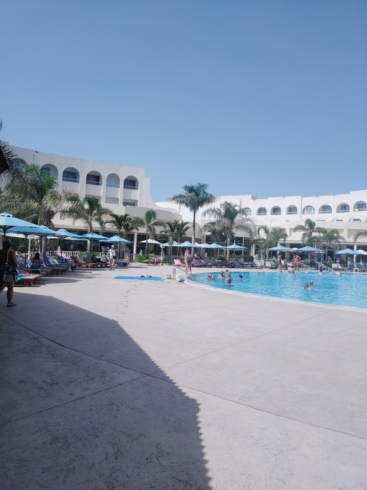 This is a wide-angle, eye-level shot of a resort swimming pool area on a clear, sunny day. The large, blue swimming pool occupies the right two-thirds of the frame, with several people visible in the water, swimming and standing. The pool area is paved with light-colored concrete. Along the left side of the pool, there is a row of blue umbrellas and lounge chairs, some occupied by people. Behind the pool area and lounge chairs is a multi-story, white hotel building with arched windows and balconies, typical of coastal resorts in Nabeul, Tunisia. Palm trees are scattered throughout the scene, adding to the tropical atmosphere. The sky is a clear, bright blue. The overall impression is that of a lively, holiday resort scene.