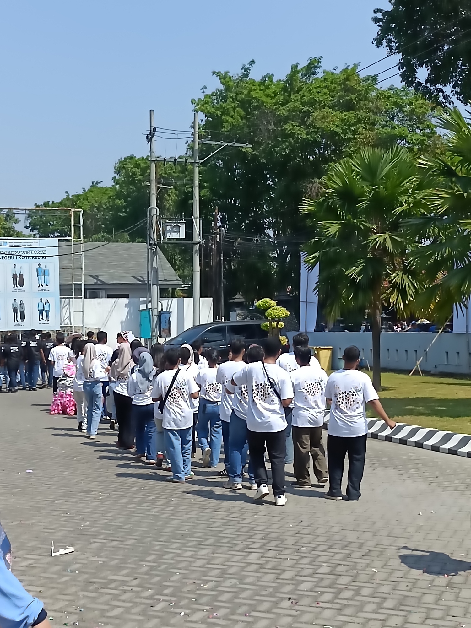 The image shows a group of people, mostly young adults, walking in a line on a paved walkway in an outdoor setting. They are all wearing white t-shirts with a circular pattern on the back and blue jeans. The people are facing away from the camera and appear to be participating in an event or procession.
In the background, there is a building with a white facade and a large banner with graphics depicting different uniforms. There are also trees, palm trees, power lines, and a black car. The walkway is made of interlocking bricks and there is some debris scattered on the ground.
The location is identified as Kediri City, Indonesia, based on the banner. The overall scene suggests a public event or gathering taking place during the daytime.