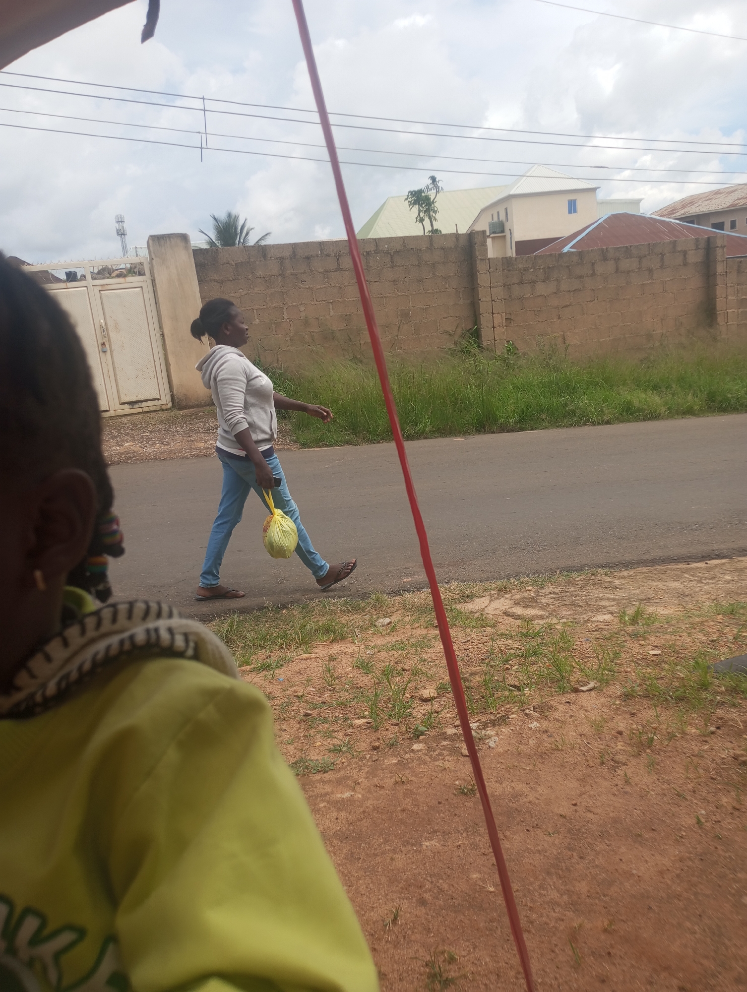 A young Black woman in a grey hoodie and blue jeans walks on an asphalt road in a city environment, possibly Jos, Nigeria. She is carrying a yellow plastic bag in her right hand. In the foreground, a child's shoulder and head are visible, partially obscuring the scene. The background features a beige brick wall, a white gate, and buildings with various roof types. The sky is cloudy. A tall red pole is positioned on the right side of the image.