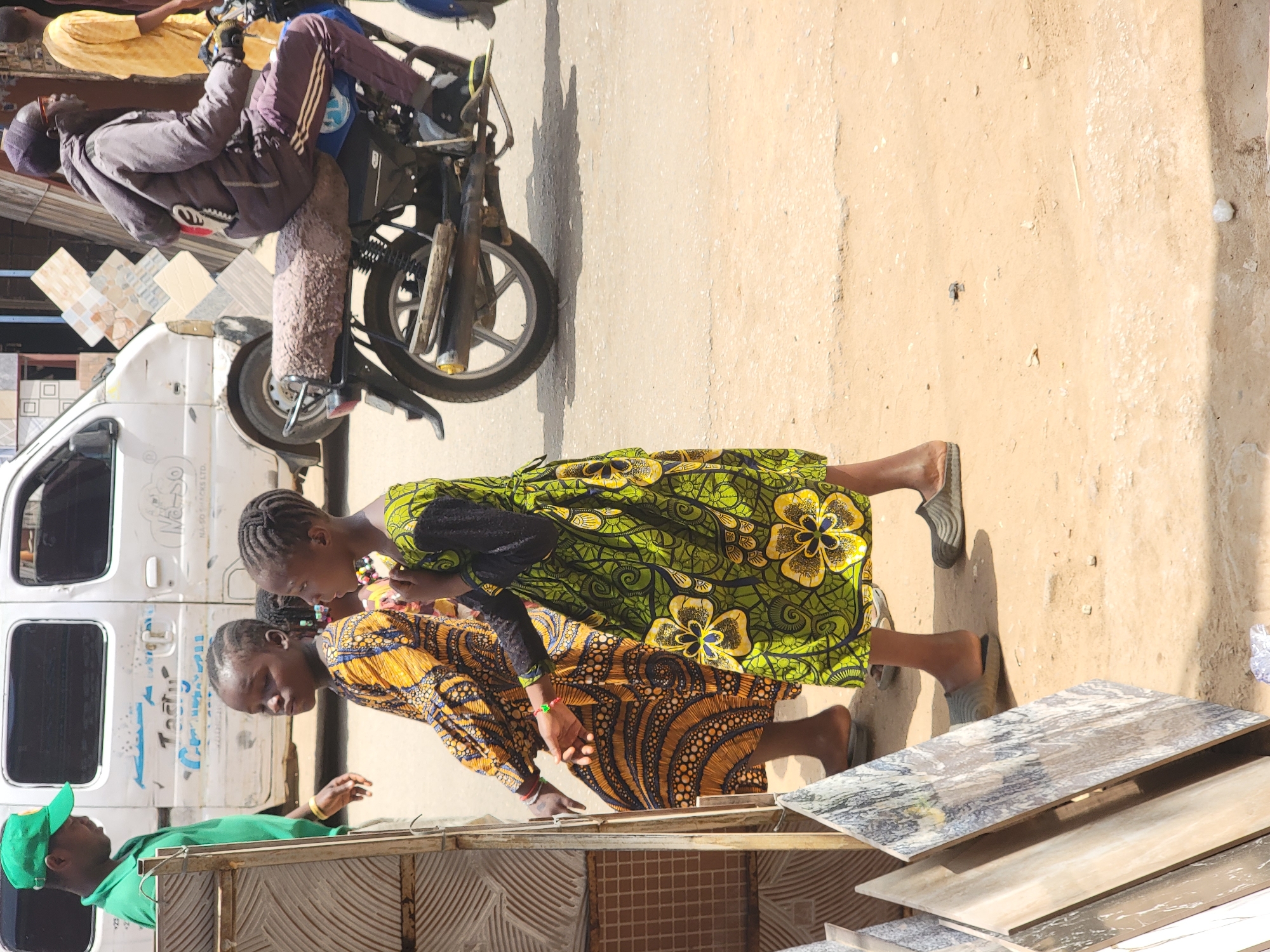 Two women, one in a green and yellow floral dress, the other in a patterned brown and yellow dress, stand beside a rack of tiles. A man in a green shirt and cap is also present. A parked white van and a motorcycle are nearby. The setting appears to be an outdoor marketplace or commercial area. This is in Maiburiji, Nigeria.