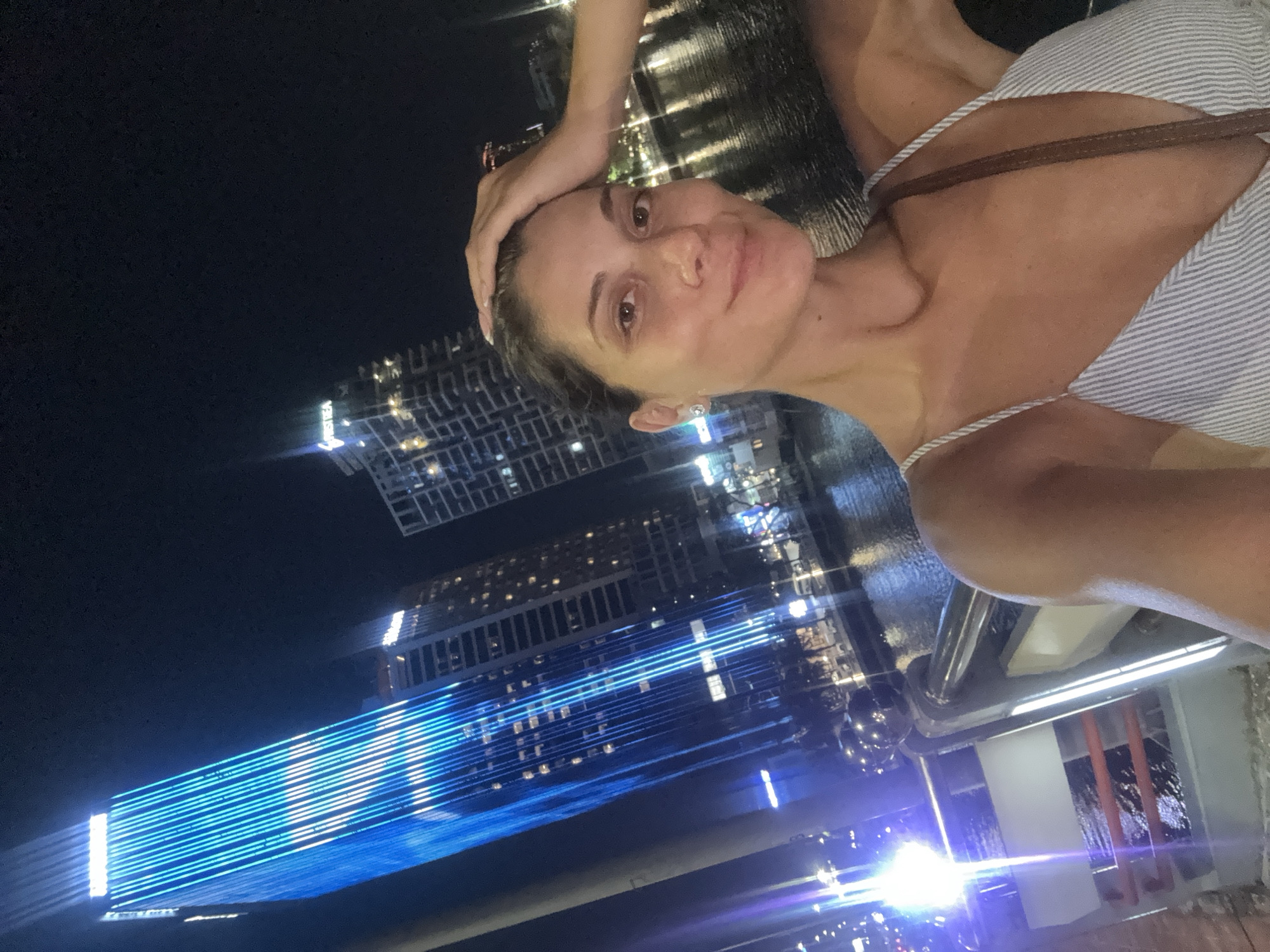 A selfie taken at night shows a woman with her right hand on her head. In the background, there are skyscrapers and water. The skyscrapers have multiple floors and are brightly lit. The water appears to be reflecting the lights of the city. The woman is wearing a light-colored top with thin straps. The setting is likely a waterfront area in the city of Đà Nẵng, Vietnam, given the presence of modern high-rise buildings and a body of water that is probably the Han River.