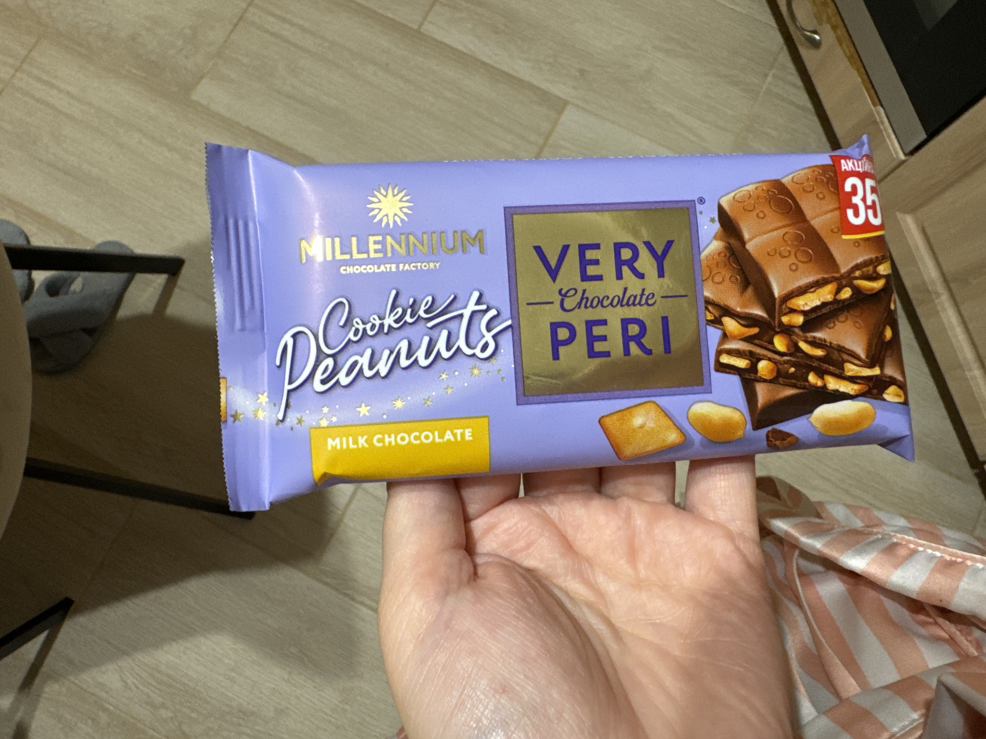 A person's left hand holds a purple chocolate bar. The chocolate bar has a yellow label that says "MILK CHOCOLATE." There is also text that says "Millennium Chocolate Factory." Below that, there is a golden square with text that says "- Chocolate VERY PERI". A picture of chocolate with peanuts is on the front of the bar. A red sticker with "35" is on the bottom right of the bar. The background appears to be a wooden floor.