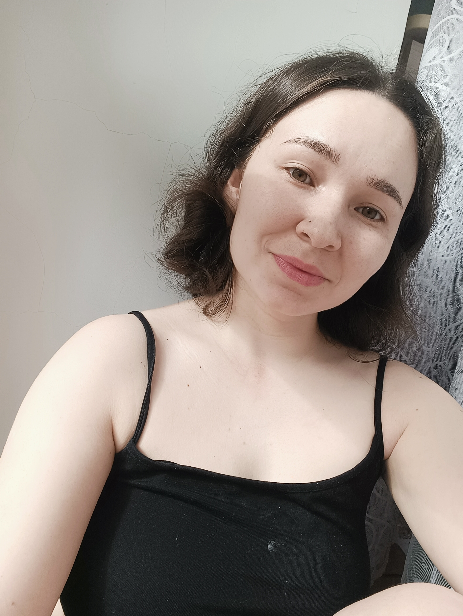 A young woman is posing for a selfie. She has short, dark brown hair and hazel eyes. She is wearing a black tank top. A nose ring is visible on the left side of her nose. The background consists of a white wall on the left and a light gray curtain on the right. The location is possibly in a residential setting, but there is no information to suggest that this image was taken in the city of Izhevsk, Russia.