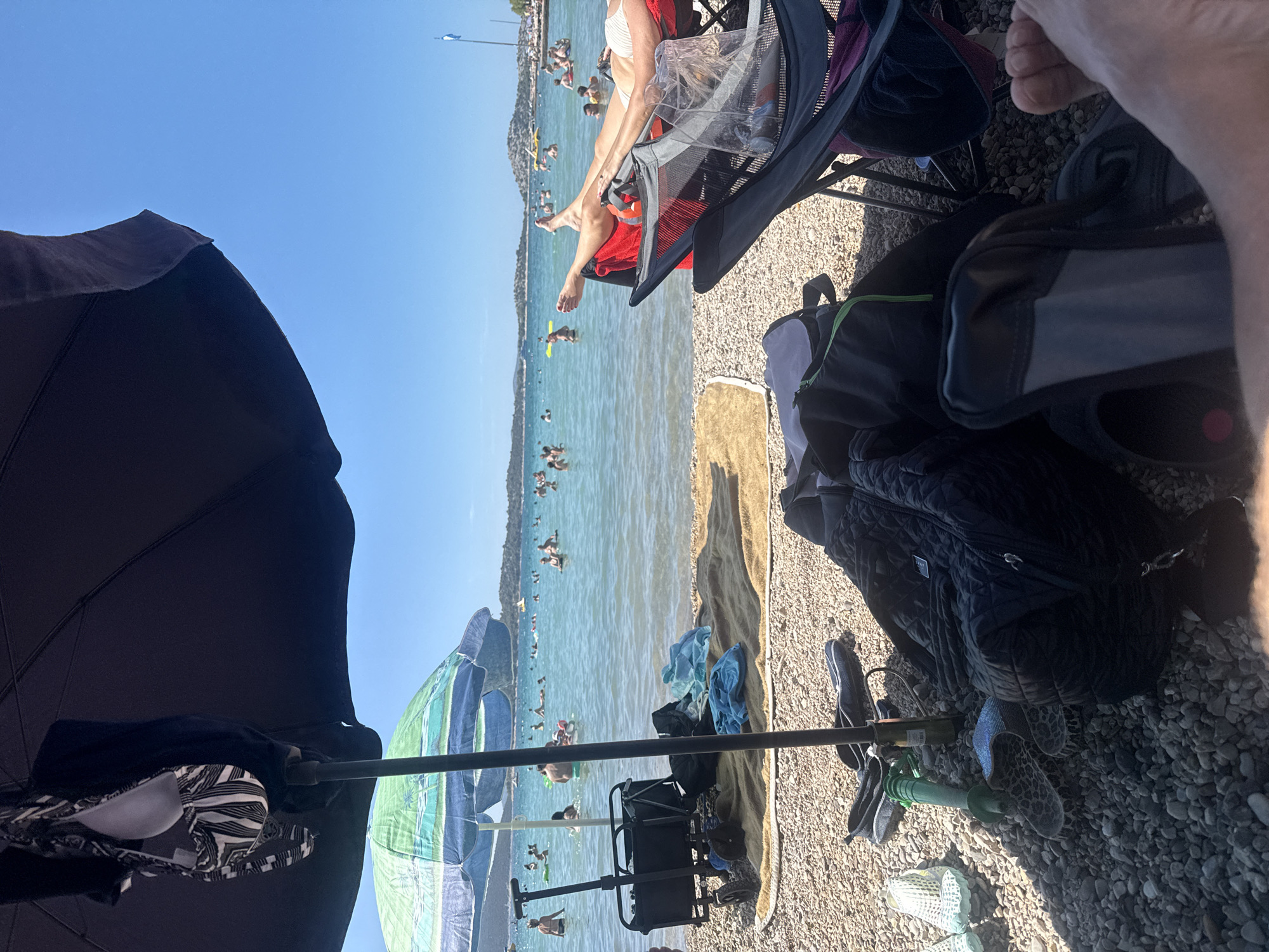 The image depicts a sunny day at a pebble beach in Vodice, Croatia, with a clear blue sky and calm turquoise water. In the foreground, several beach accessories are laid out on the pebbles. These include a black foldable chair with a person's legs resting on it, a large black duffel bag, a smaller black backpack with a green zipper, a green water hose with a nozzle, a pair of patterned slide sandals, and a pair of light-colored water shoes. A large dark umbrella shades a portion of the scene, and a colorful beach umbrella is also visible. In the middle ground, a black beach cart is parked on the pebbles. The water is populated with numerous people swimming, floating on inflatable toys, and enjoying the sea. In the background, a coastline with buildings and vegetation is visible, with several islands or peninsulas in the distance. A flag on a pole is also present on the right side of the frame. The overall impression is one of a relaxed and enjoyable beach day.