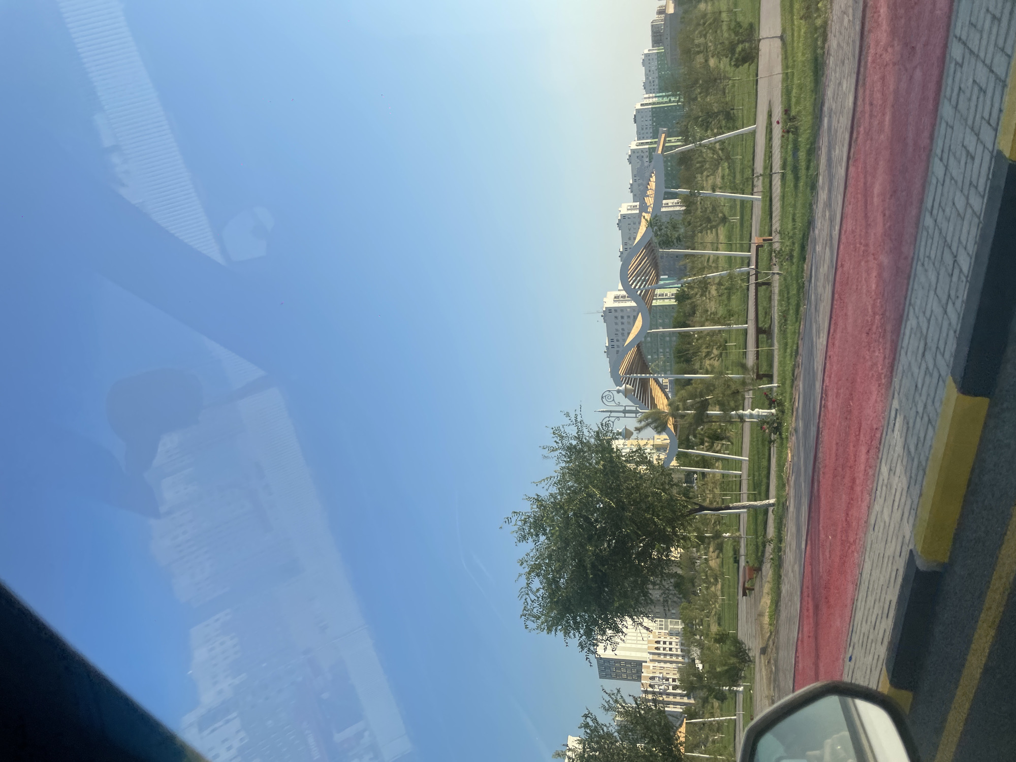 The image depicts a view of a modern park in the city of 30 Years of Kazakhstan, Kazakhstan, as seen from a vehicle. In the foreground, a red jogging path runs alongside a gray and yellow brick sidewalk. The park itself features lush green grass, scattered trees, and stylized white and wooden covered seating areas. Beyond the park, a line of tall, modern apartment buildings with light-colored facades stretches across the horizon under a clear blue sky. Reflections of clouds and possibly parts of the vehicle's interior are visible on the windshield.