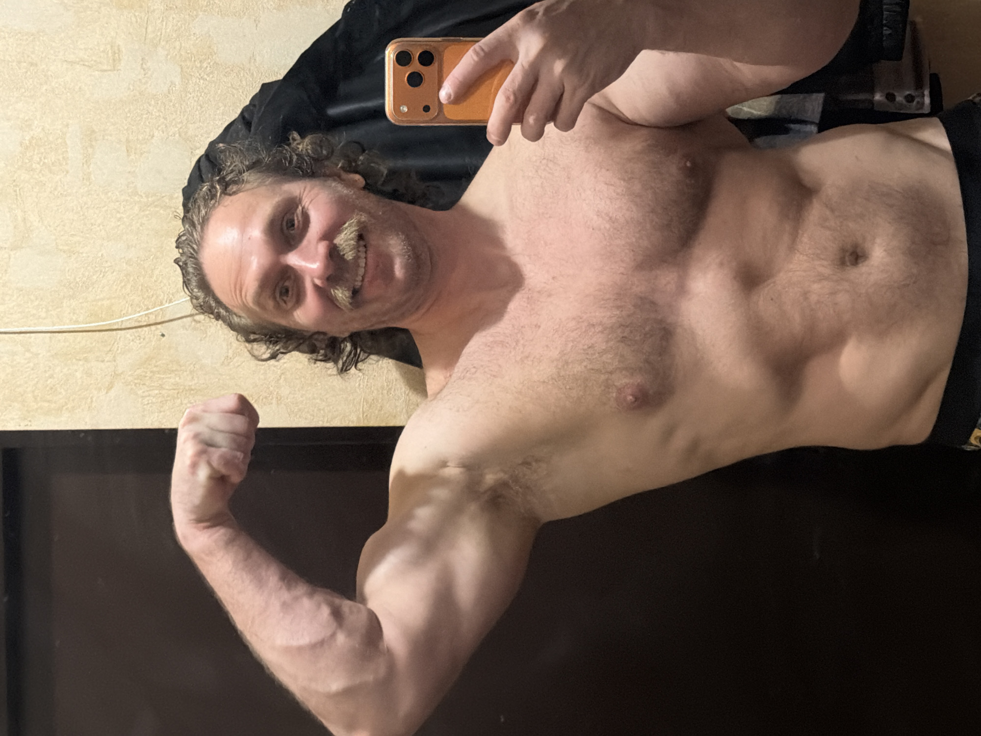 The image displays a shirtless man taking a selfie indoors. He is positioned with his head towards the upper left and his body extending diagonally across the frame. The man has curly dark hair and a prominent light-colored mustache, and he is smiling broadly at the camera, conveying a sense of pride or contentment. His right hand is holding a smartphone, which has a distinctive gold or orange case and a triple-lens camera array, suggesting it might be an iPhone.
He exhibits a muscular and toned physique, with visible chest muscles, defined abdominal areas, and a well-developed left arm which he is flexing to showcase his bicep. There is visible body hair on his chest and abdomen. The background features a light-colored, textured wall, possibly stucco, with a white cable running along it. A black garment or fabric is draped over his left shoulder, and a large dark, reflective surface, likely a television screen or furniture, occupies the lower right portion of the frame. The lighting in the room appears to be artificial and warm. The image's location context is Saint Petersburg, Russia.