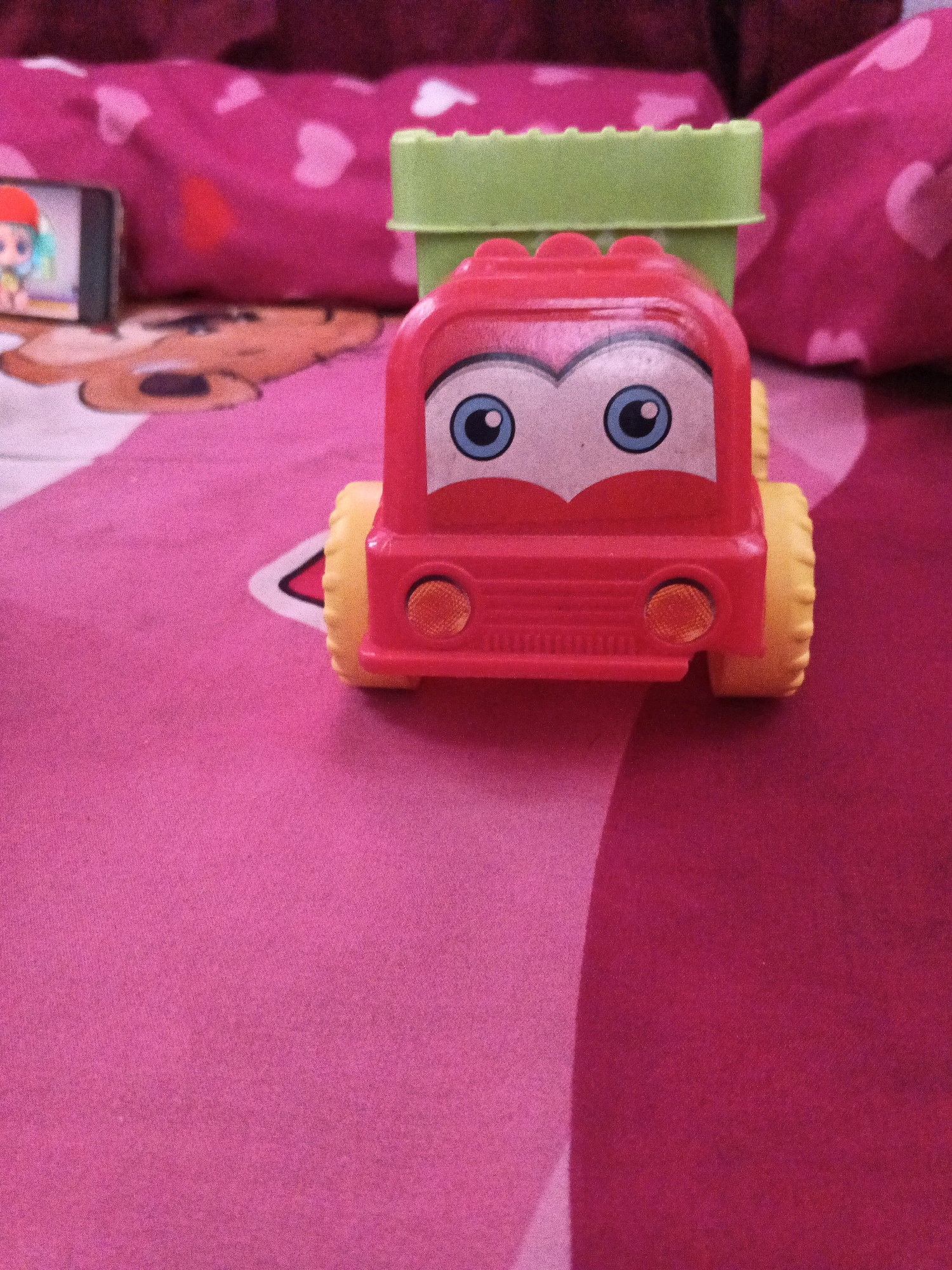 A red toy truck with a green bed sits on a pink surface with a teddy bear pattern. The truck has large blue eyes and yellow wheels. In the background, there is a pink pillow with white heart shapes and a smartphone displaying a cartoon character. The scene is indoors. The city of Gabus, Indonesia, is not visible in this image.