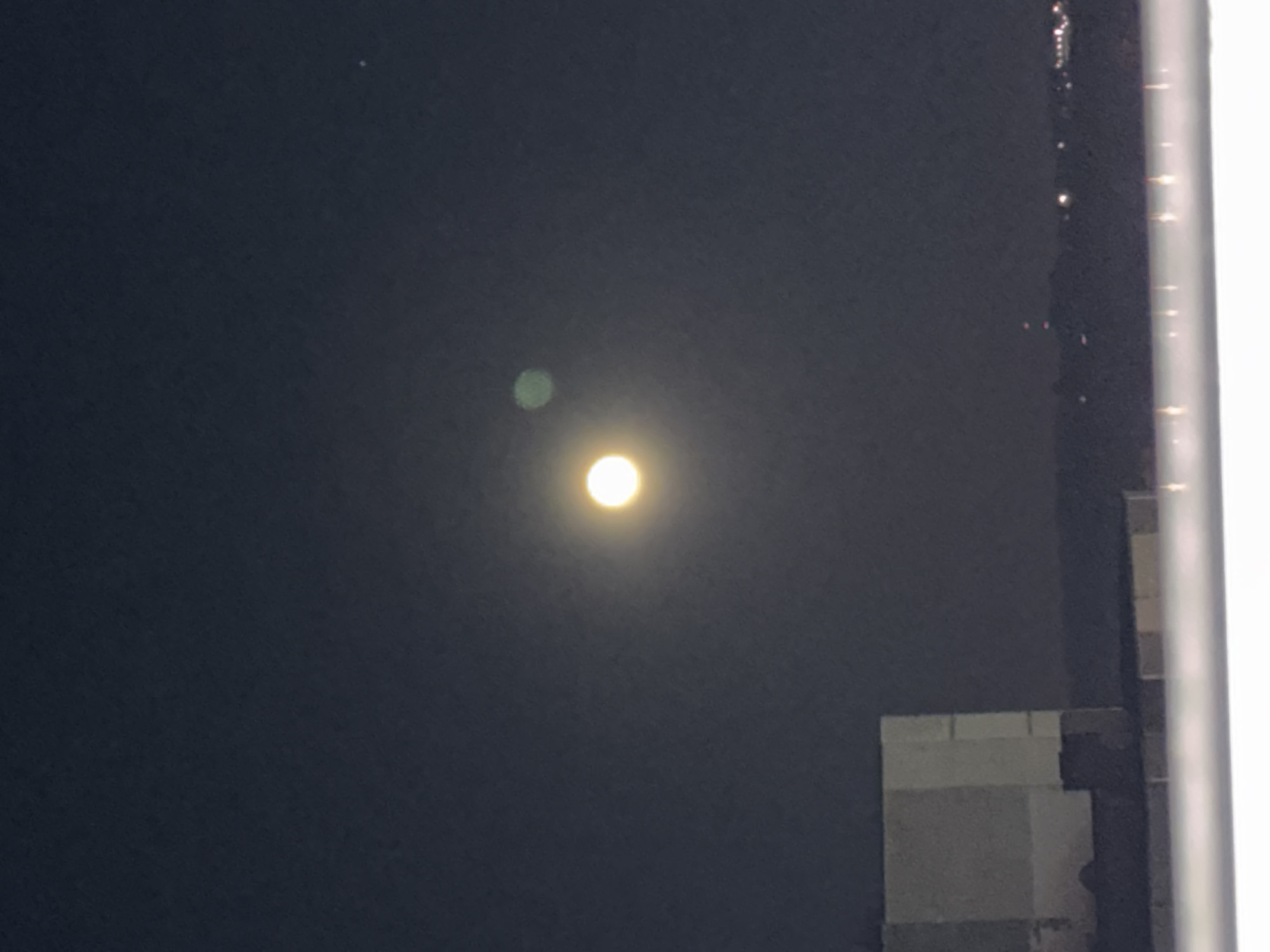 The image shows a night scene with a bright, spherical object, likely the moon, in the dark sky. A smaller, green circular shape is faintly visible to the upper left of the brighter object. In the lower right, parts of a building are visible, including some white or light-colored walls or structure. In the far right, a white, vertical line possibly a window frame, is seen. The location of Minsk, Belarus is not discernible from the visual information.