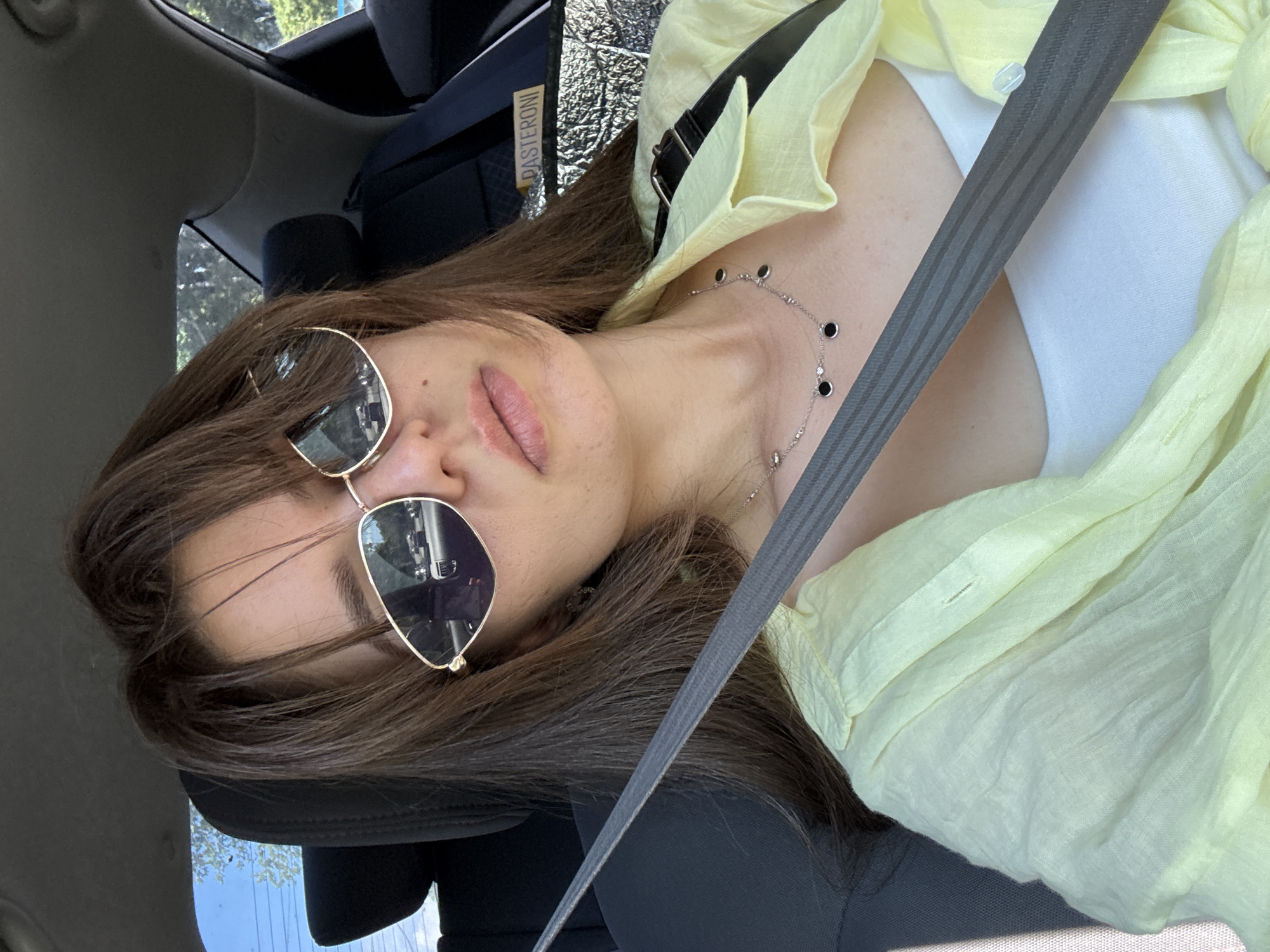 A young woman with long brown hair and sunglasses is taking a selfie in a car. She is wearing a light yellow button-up shirt over a white tank top and a silver necklace with black and clear stones. A grey seatbelt is diagonally across her chest. The interior of the car is visible, including the headrest of the passenger seat and part of the dashboard. Outside the car, trees and a blue sky can be seen. A sign with the word "PASTERONI" is visible on the seat behind her.