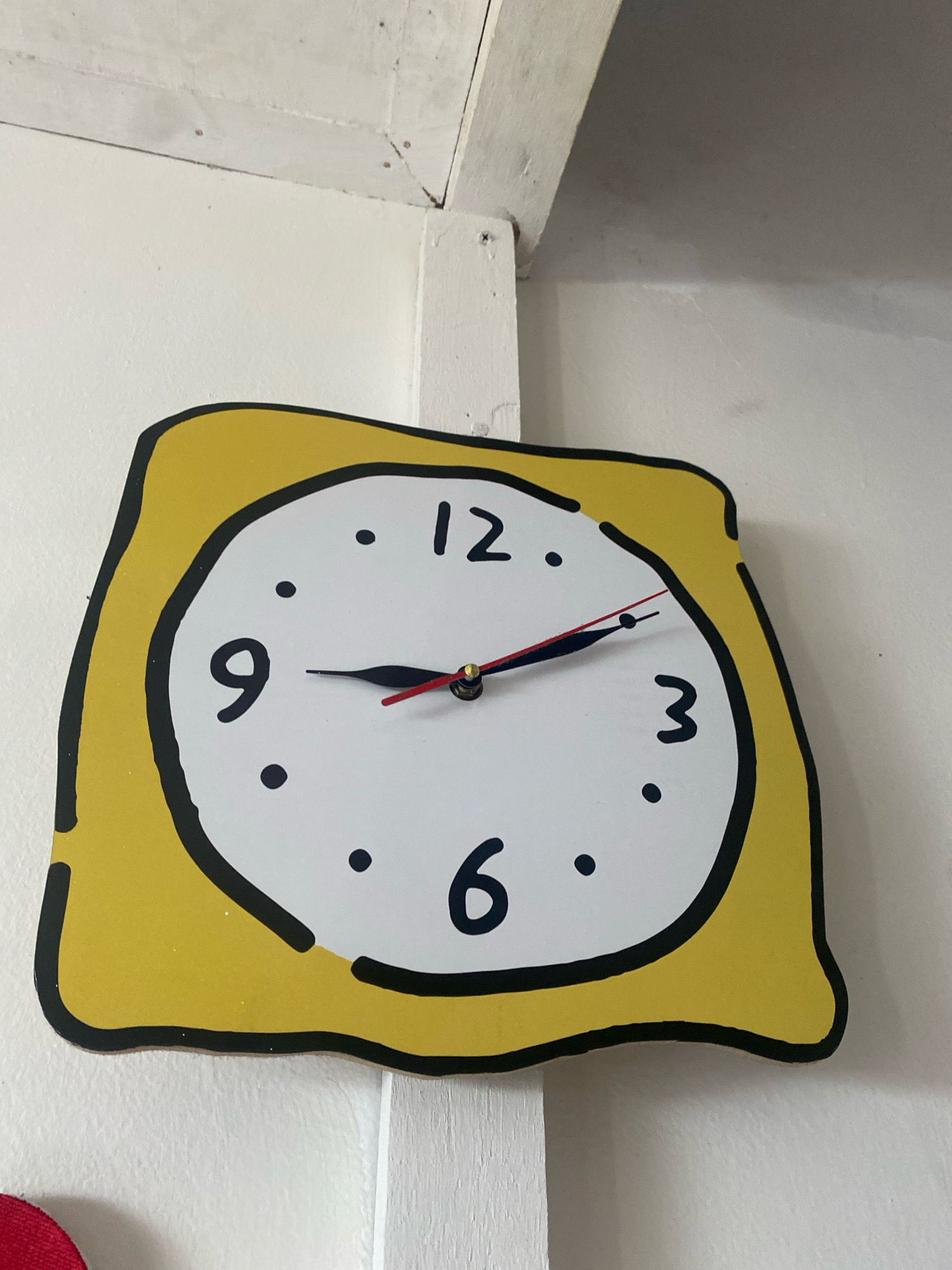A close-up, low-angle, eye-level shot shows a yellow, irregularly shaped clock with a black outline hanging on a white wall. The clock face is white with black numbers indicating the hours from 1 to 12, and black dots marking the minutes. Two hands are visible: a shorter, thicker black hand pointing between 9 and 10, and a longer, thinner red-tipped black hand pointing slightly past the 1. The clock is mounted on a white wall, with visible white wooden beams at the top of the frame. This image was captured in Bandar Lampung, Indonesia.
