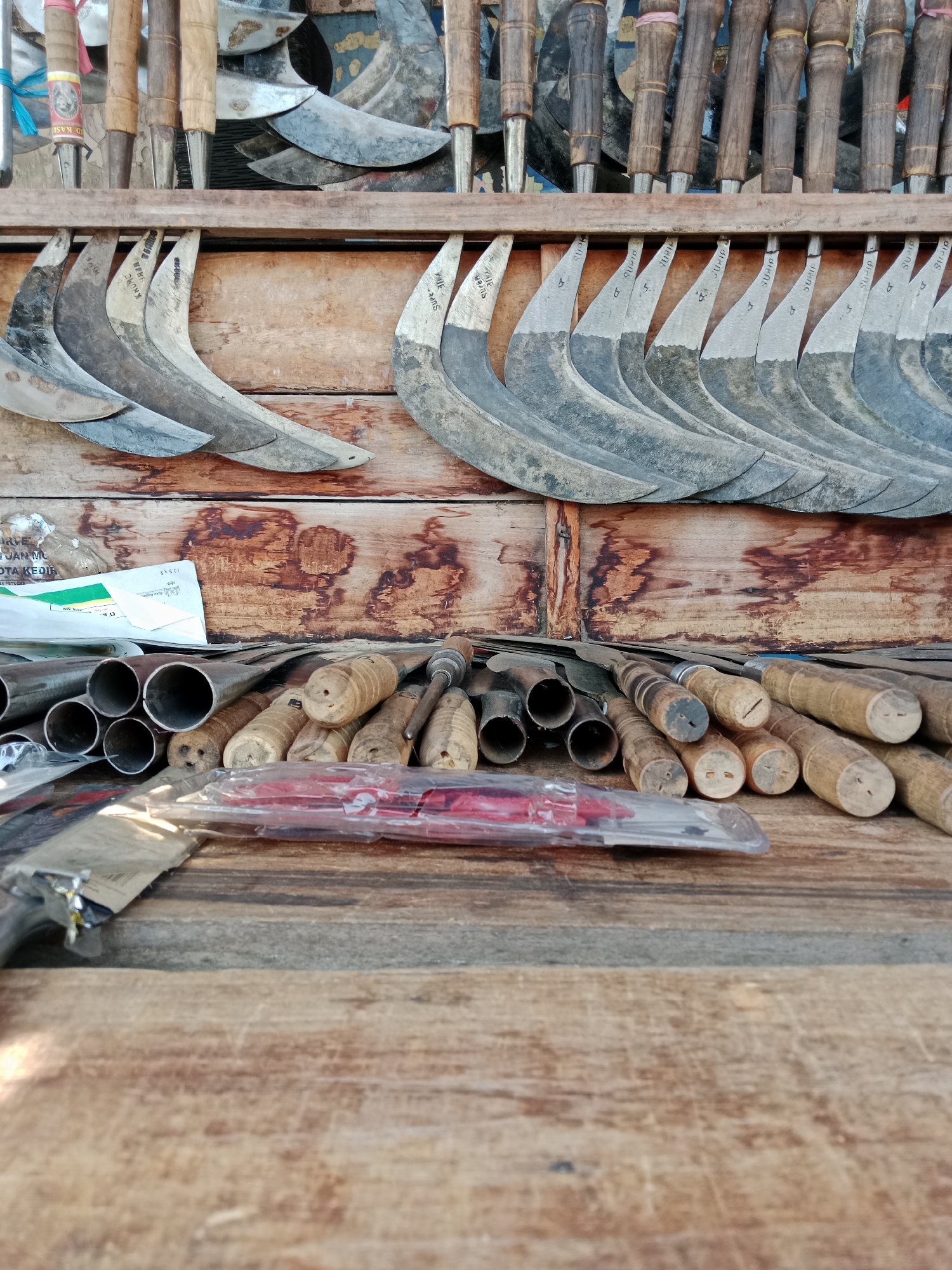 The image depicts a close-up, low-angle view of various agricultural tools and their components displayed on a wooden surface. The primary subjects are rows of sickles with curved blades and wooden handles, arranged on a wooden plank. Above these, another shelf displays more sickle blades, some with handles attached. To the left and in the foreground, a collection of metal pipes and more wooden handles are visible, along with some packaged items. The background consists of weathered wooden planks, suggesting an outdoor or rustic setting. A partially visible label on a piece of paper in the background indicates "KOTA KEDIRI," which identifies the location as Kediri City, Indonesia. The overall impression is one of a tool stall or workshop, emphasizing the craftsmanship and variety of tools, likely for farming or gardening.