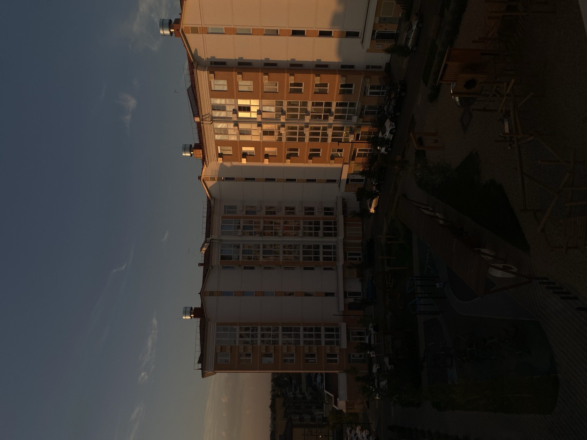 The image shows a residential building in the city of Vishnyaki, Russia, at dusk. The building is modern with multiple stories and a light-colored facade accented with orange details around the windows and roof. The windows are large and reflect the warm, golden light of the setting sun. Several cars are parked on the street in front of the building. A playground area with sand and play structures is visible in the foreground, with a pathway leading through it. The sky above is a deep blue, transitioning to a lighter orange hue near the horizon, with a few wispy clouds. The overall scene depicts a peaceful urban environment during the evening hours.
