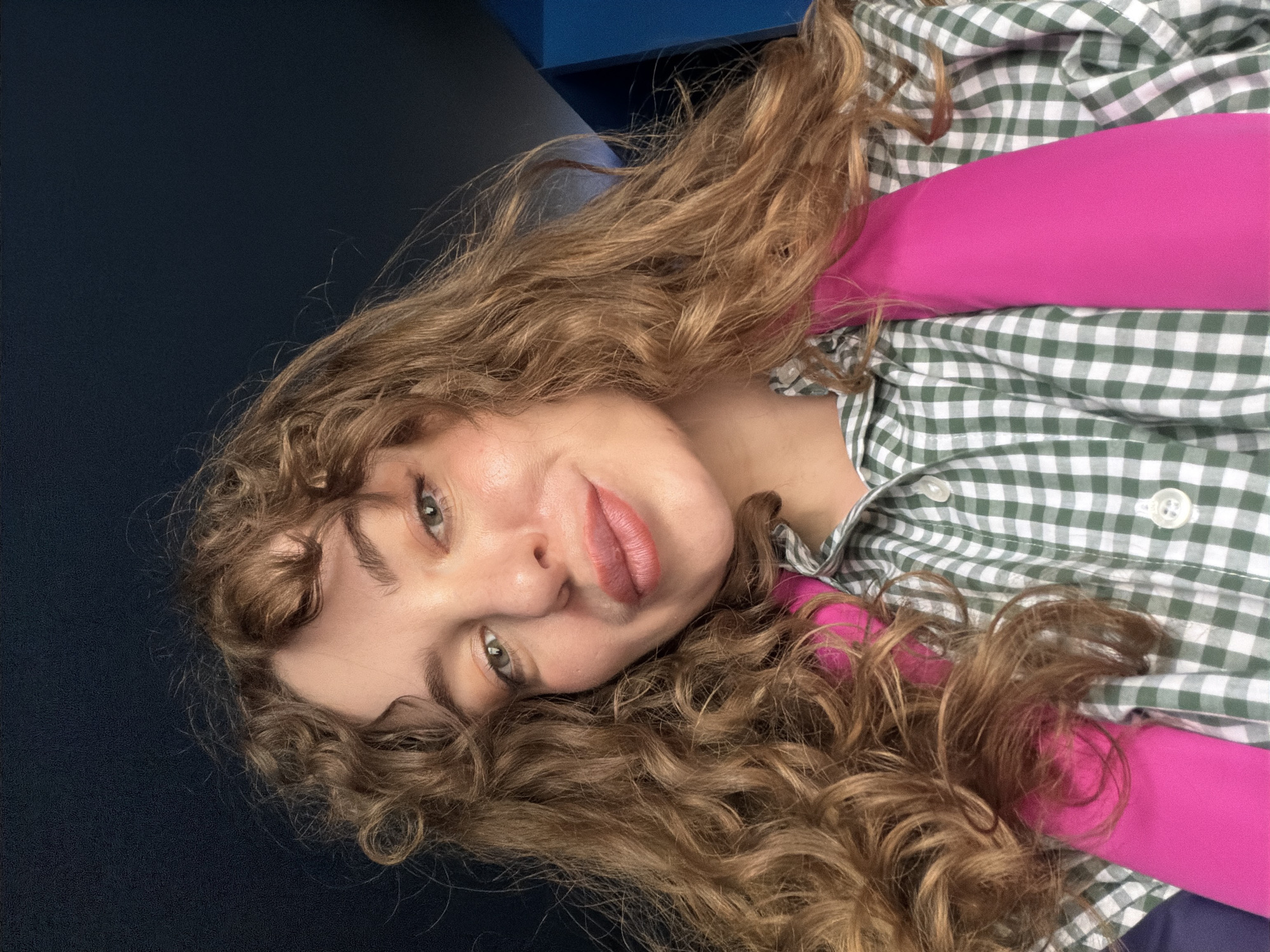 A woman with long, curly, light brown hair, fair skin, and light-colored eyes is lying on a dark blue surface. She is wearing a green and white checkered button-down shirt over a bright pink top. The background is blurred, but hints of a blue wall can be seen. There is no indication of a location in Moscow, Russia. The image is a close-up selfie or portrait.