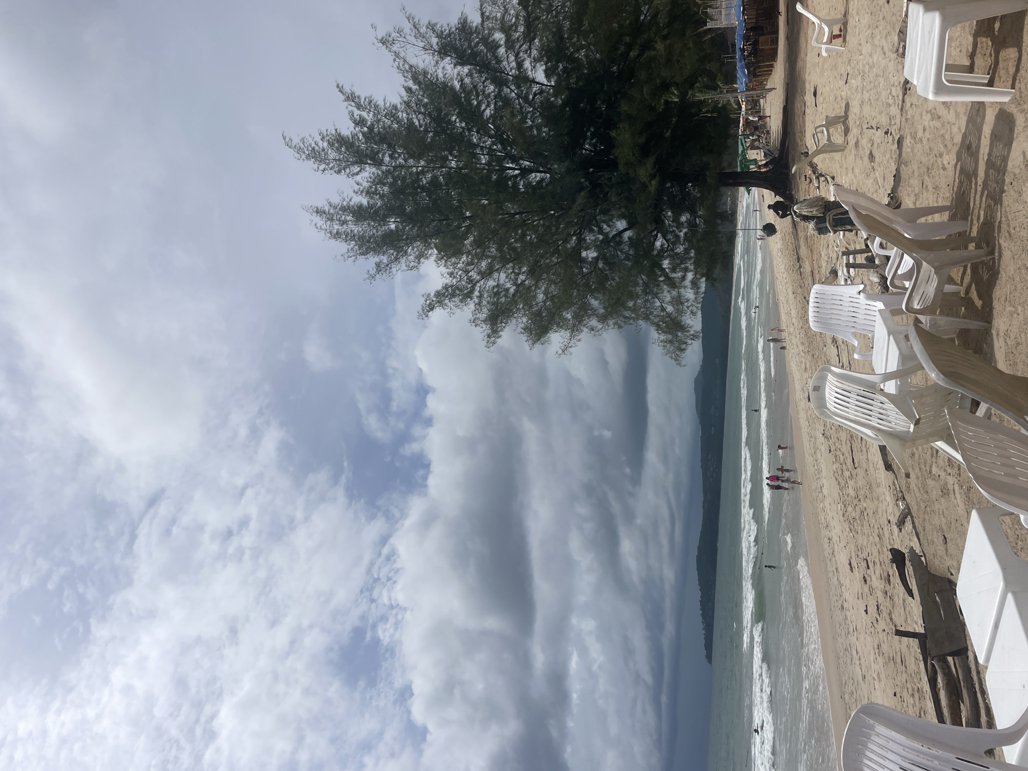 The image depicts a beach scene in Choeng Thale, Thailand. A large tree stands on the shore, near numerous white plastic chairs and tables, likely for beachgoers. The beach itself has a light tan sand, with small waves approaching the shore. Some people are walking on the beach and in the water. The sky is cloudy, with both dark and light gray clouds covering most of the visible area. The image is oriented vertically.