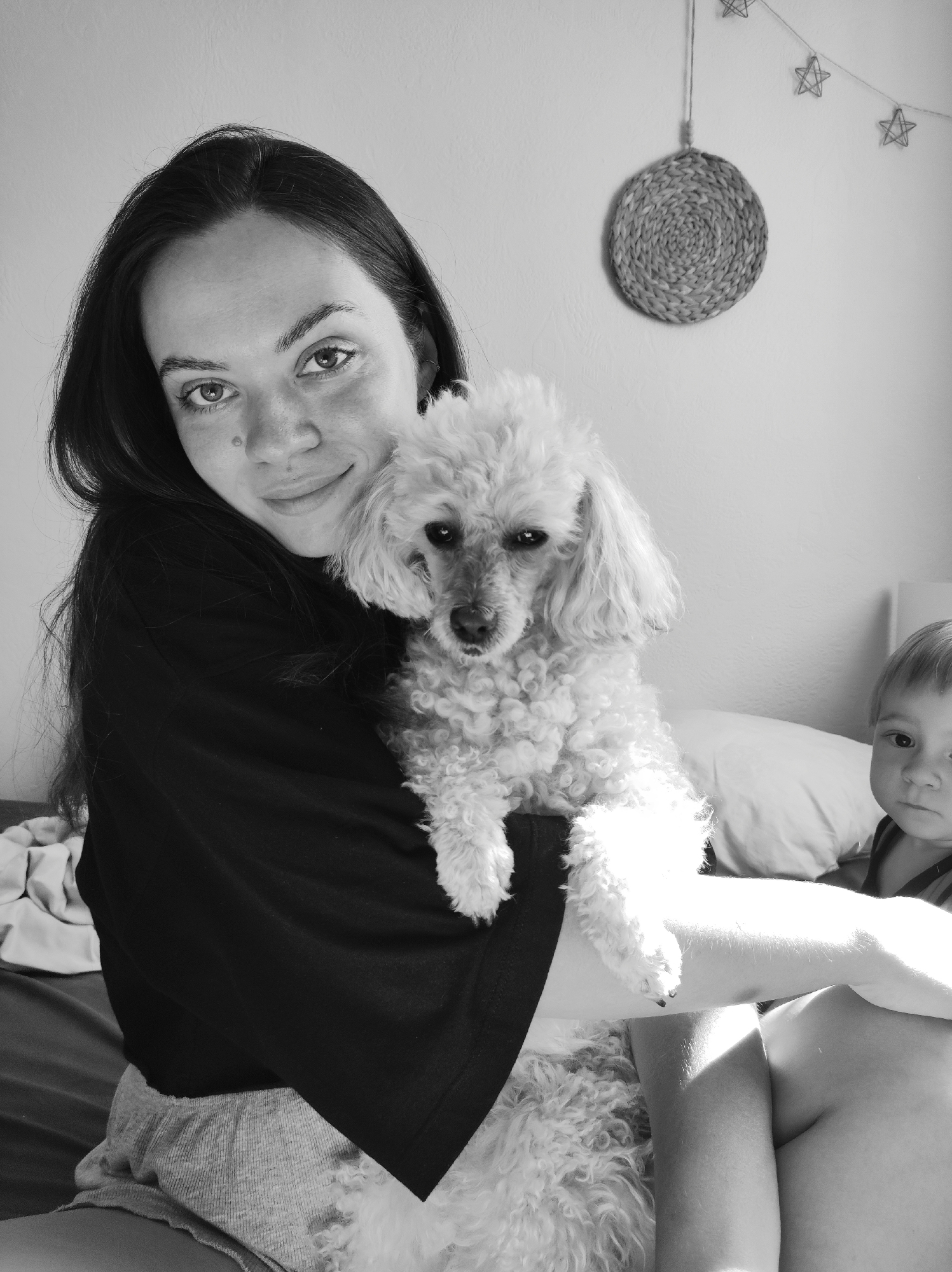 The image shows a woman holding a small, light-colored poodle dog. The woman has dark hair and is wearing a dark t-shirt. She is looking at the camera and smiling slightly. The dog is also looking at the camera. In the background, there is a decorative woven circular object hanging on the wall, a string of star decorations, a pillow, and a young child with short blond hair partially visible on the right side of the image. The scene appears to be indoors, possibly in a bedroom. There is no visual evidence to confirm the location being in Chaikovsky, Russia.