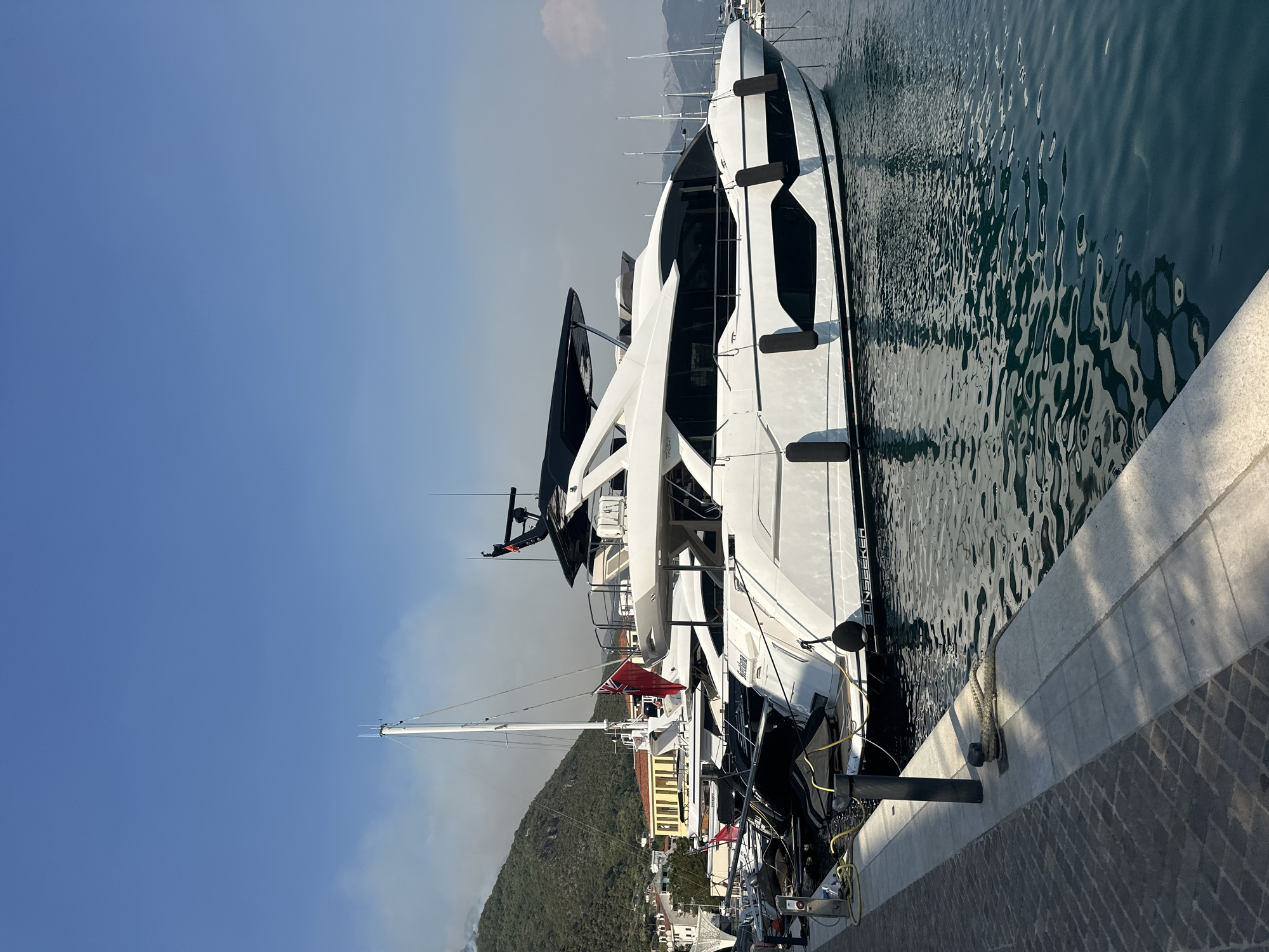 A modern, white motor yacht with black accents is moored at a marina in Herceg Novi, Montenegro. The yacht, identified by the name "SUNSEEKER" on its hull, is sleek and features multiple levels and tinted windows. It is secured to a paved walkway by thick ropes and black fenders. Other sailboats and smaller vessels are visible in the background, along with a tall white mast. The marina is situated against a backdrop of a green, hilly landscape with scattered buildings. The sky is clear blue, with some wisps of smoke visible in the distance, suggesting a potential fire on the hillside. The water is calm, with reflections of the yachts visible on its surface. The lighting indicates it is daytime, likely morning or afternoon, with bright sunlight casting shadows.