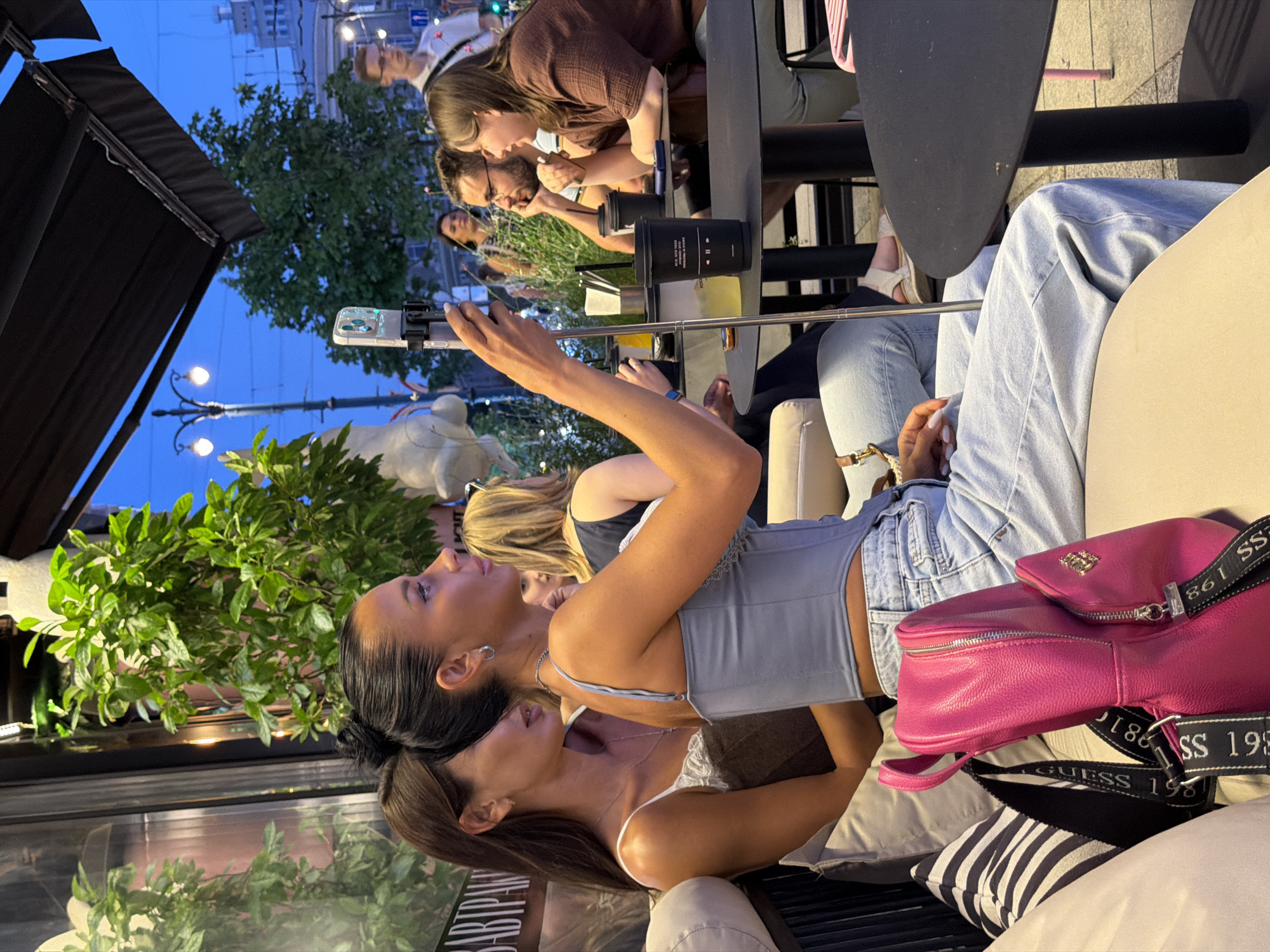 The image depicts a group of people seated at outdoor tables in a cafe setting, possibly in Novaya Usman', Russia, given the context of the prompt. In the foreground, a woman is holding a selfie stick with a smartphone attached, angled towards herself and another woman seated next to her. Both women are wearing casual attire. The woman holding the selfie stick has dark hair pulled back in a bun and is wearing a light blue corset-like top and light-wash jeans. The woman next to her has long brown hair and is wearing a white top with lace trim. To their right, another woman with blonde hair is visible, and further in the background, a man with glasses and a woman are engaged in conversation. A bright pink backpack rests on the seating area in the foreground. The environment appears to be an urban outdoor space, with trees, streetlights, and buildings visible in the background, suggesting a city location. The lighting indicates it is either dusk or early evening.