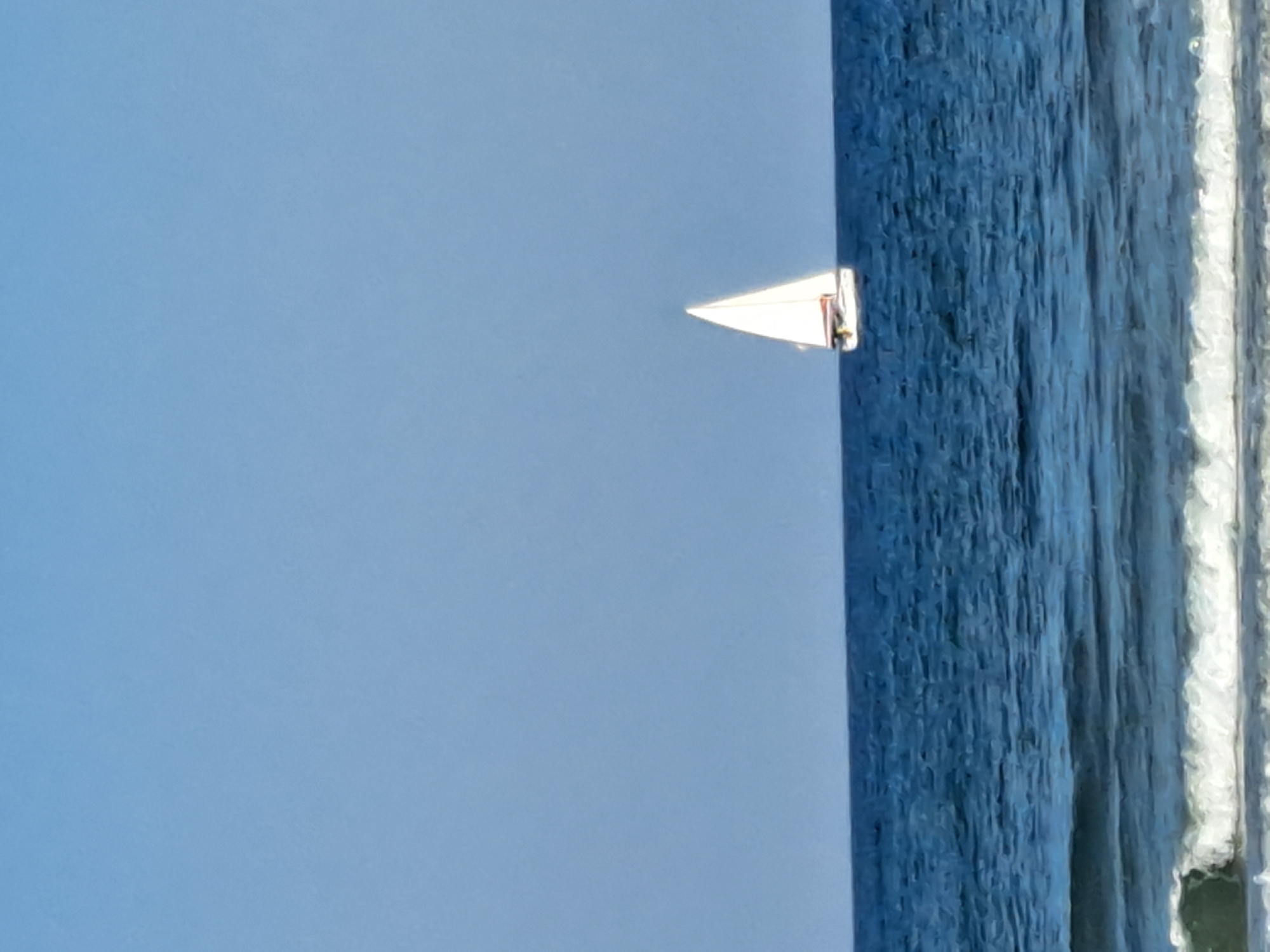 The image depicts a serene maritime scene under a clear blue sky, likely taken from the coastline of Herzliya, Israel. The vast expanse of the deep blue Mediterranean Sea dominates the lower half of the frame, with gentle waves cresting white in the foreground, indicating a shallow shoreline. Further out, a single white sailboat with a red stripe along its hull is visible, its sail taut against the breeze, sailing gracefully on the water. The horizon line is sharp, separating the sea from the cloudless sky. There are no visible people or structures in the immediate vicinity, emphasizing the tranquil and natural beauty of the location.