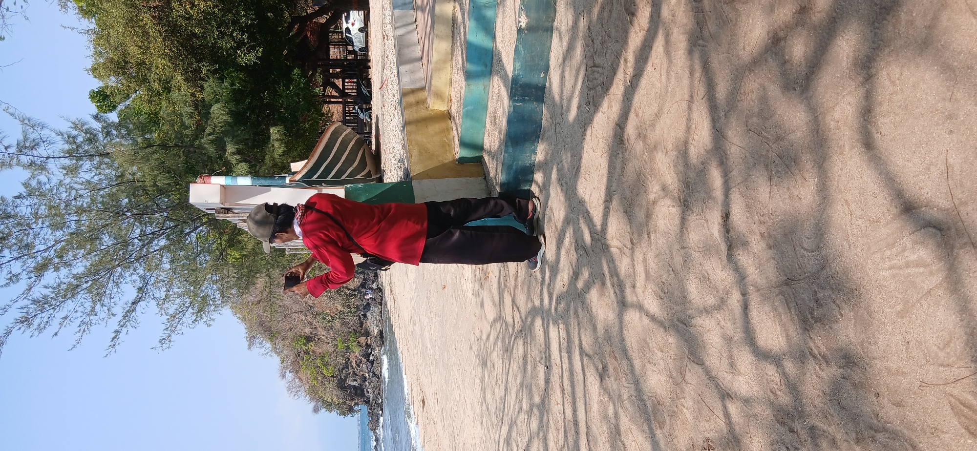 A person wearing a red long-sleeved shirt, dark pants, a green cap, and a black crossbody bag stands on a sandy beach in Banyuglugur, Indonesia. The person is holding a smartphone in their hands, seemingly taking a picture or recording a video. The background shows a body of water, a rocky cliff, trees, and a boat. There are also some steps painted in blue and yellow on the right side of the image. The ground is covered in sand with tree shadows cast upon it.