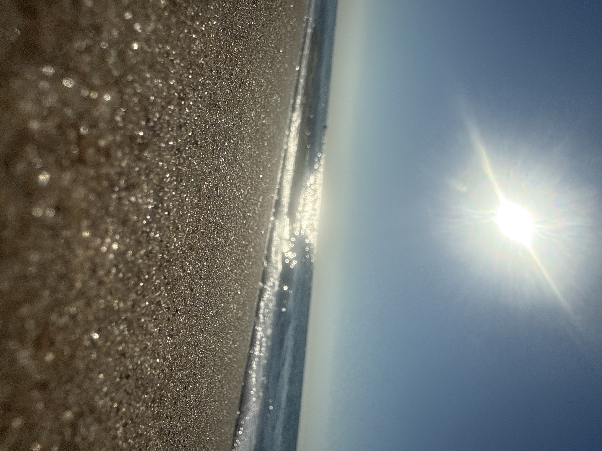 The image depicts a serene beach scene under a bright sun. The foreground is dominated by the shimmering sand, with individual grains glinting under the light. A gentle wave is rolling in, creating a foamy white edge along the shoreline. The ocean stretches out to the horizon, reflecting the sunlight in a dazzling display. The sky is a clear, soft blue, with the sun positioned high and radiating bright rays. There are no visible people or man-made structures, suggesting a natural and unpopulated environment at the beach in Zatoka, Ukraine.