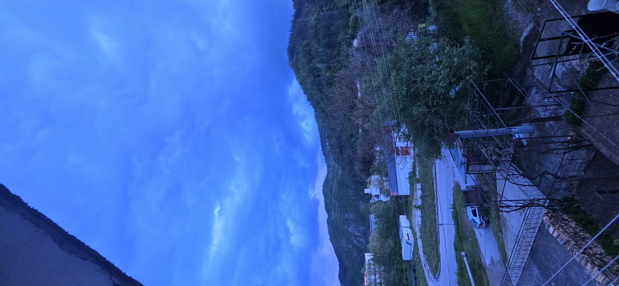 The image depicts an evening view in Rudozem, Bulgaria. A densely wooded mountain rises in the background, with the lower slopes dotted with residential buildings. The foreground shows an elevated perspective, possibly from a balcony. Visible are a paved road with street lamps, residential houses, and parked vehicles. Architectural elements like wrought iron fences and building structures are also visible. The sky is overcast, casting a blue hue over the scene.