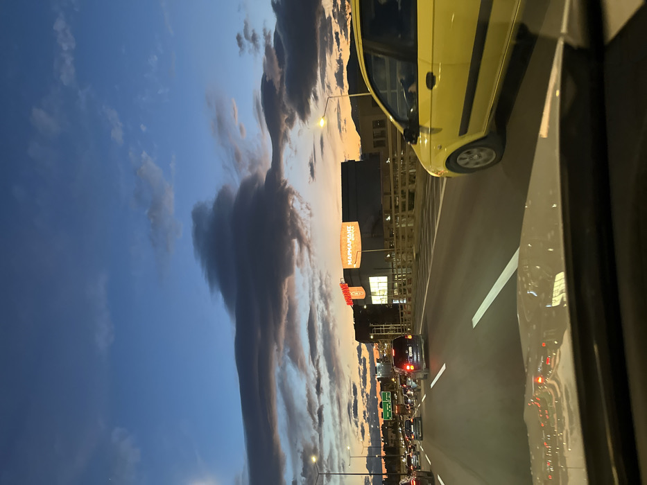 This image, oriented in landscape, captures an urban evening or morning scene from the perspective of a moving vehicle. The sky dominates the upper and left parts of the frame, showcasing a striking gradient from deep blue at the top to warm orange and pink hues near the horizon, characteristic of either dawn or dusk. Dramatic, layered clouds, some appearing dark and heavy, others lighter and more dispersed, stretch across the sky, creating a dynamic visual element.
In the mid-ground, a multi-lane road is busy with traffic. A bright yellow car, possibly a taxi, is prominently featured on the right side of the road, moving in the same direction as the viewer. The immediate foreground reveals the side window and interior reflections of the vehicle from which the photograph was taken, showing mirrored lights and road details. Further down the road, numerous vehicles can be seen, their headlights and taillights glowing, indicating significant traffic congestion.
Along the left side of the road, urban buildings and streetlights are visible. A large, orange commercial sign stands out, displaying the text "ΜΑΡΚΑΡΙΑΝΤ BROTHERS". Below it, another sign with red lettering reads "ΚΑΡΠΟΣ". The combination of natural twilight colors and artificial city lights illuminates the scene. The image conveys the atmosphere of a bustling city like Peristeri, Greece, during a transitional period of the day, with a focus on the interplay between the urban environment and a picturesque sky.