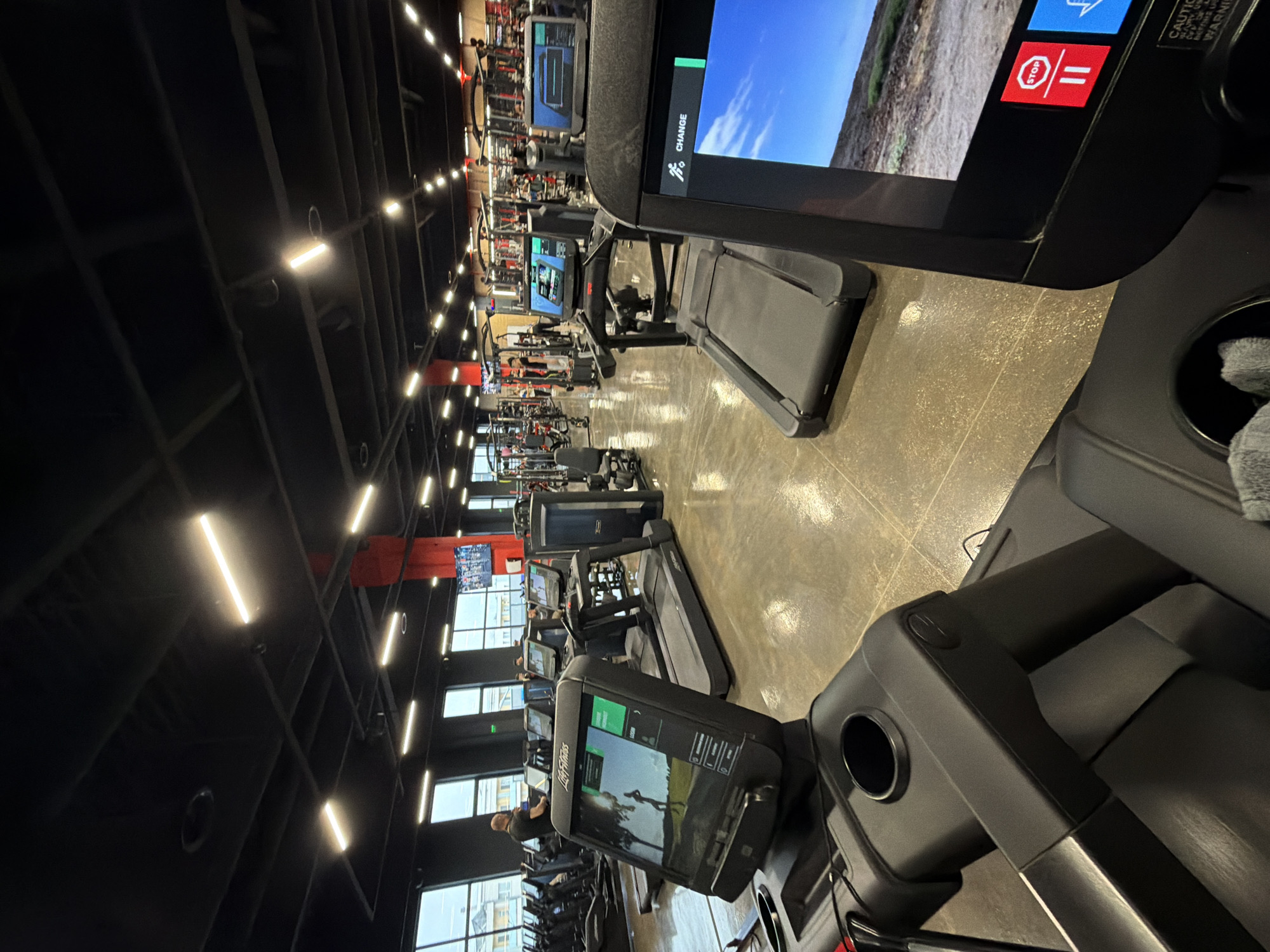 The image shows the interior of a gym with rows of treadmills. The treadmills are modern, with large screens. The gym has a dark ceiling with rows of lights. Other exercise equipment and people exercising can be seen in the background. The location is likely not in the city of Plovdiv, Bulgaria, as the gym appears to be a chain gym common in North America.