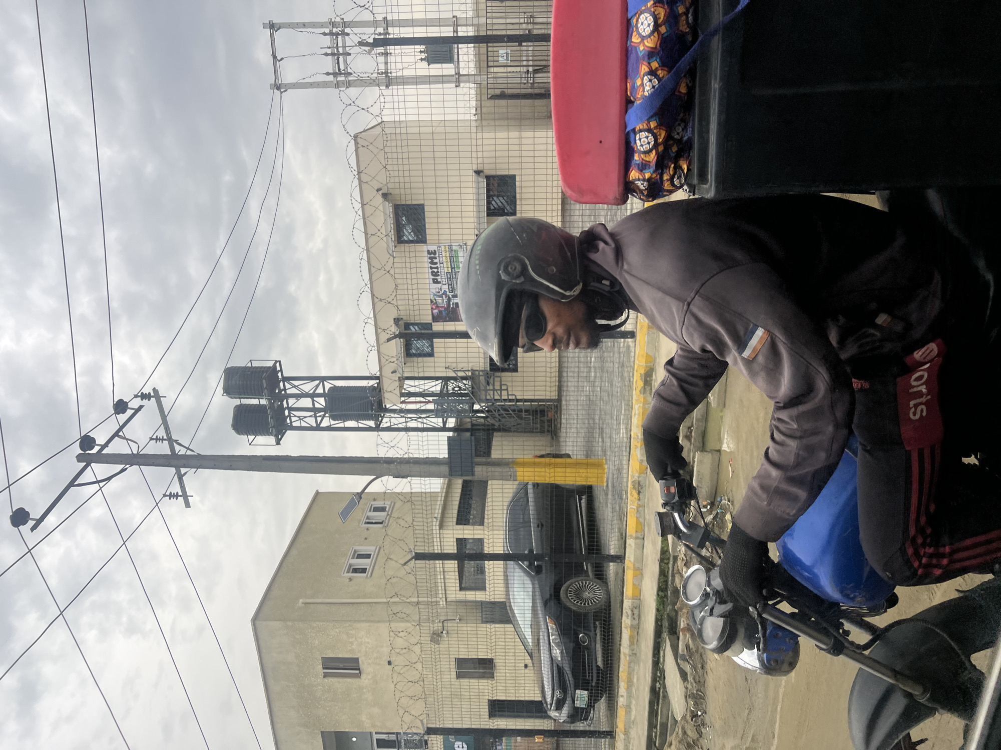 A man wearing a helmet and sunglasses is on a motorcycle in Ado, Nigeria. He is wearing gloves and a long-sleeved jacket, and pants with red stripes. The motorcycle has a blue fuel tank. Behind him is a red and black structure. In the background are buildings with barbed wire fences, a utility pole with electrical wires, water tanks on a metal support structure, and a dark-colored car parked near a yellow curb. The sky is overcast.