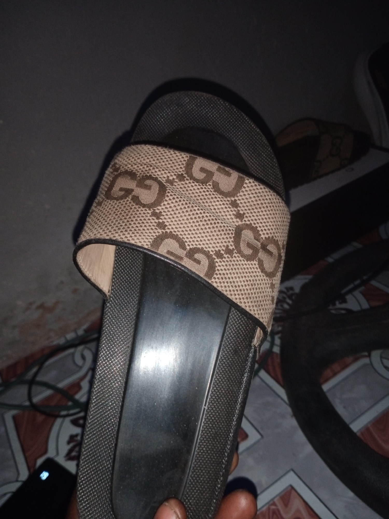 The image shows a close-up view of a person's hand holding a Gucci slide sandal. The sandal has a brown fabric strap with the iconic interlocking GG logo in a darker brown. The sole of the sandal is black and textured. The background is dark and indistinct, but appears to be an indoor setting with some tiling on the floor. There is no identifiable information about the location in Jalingo, Nigeria.