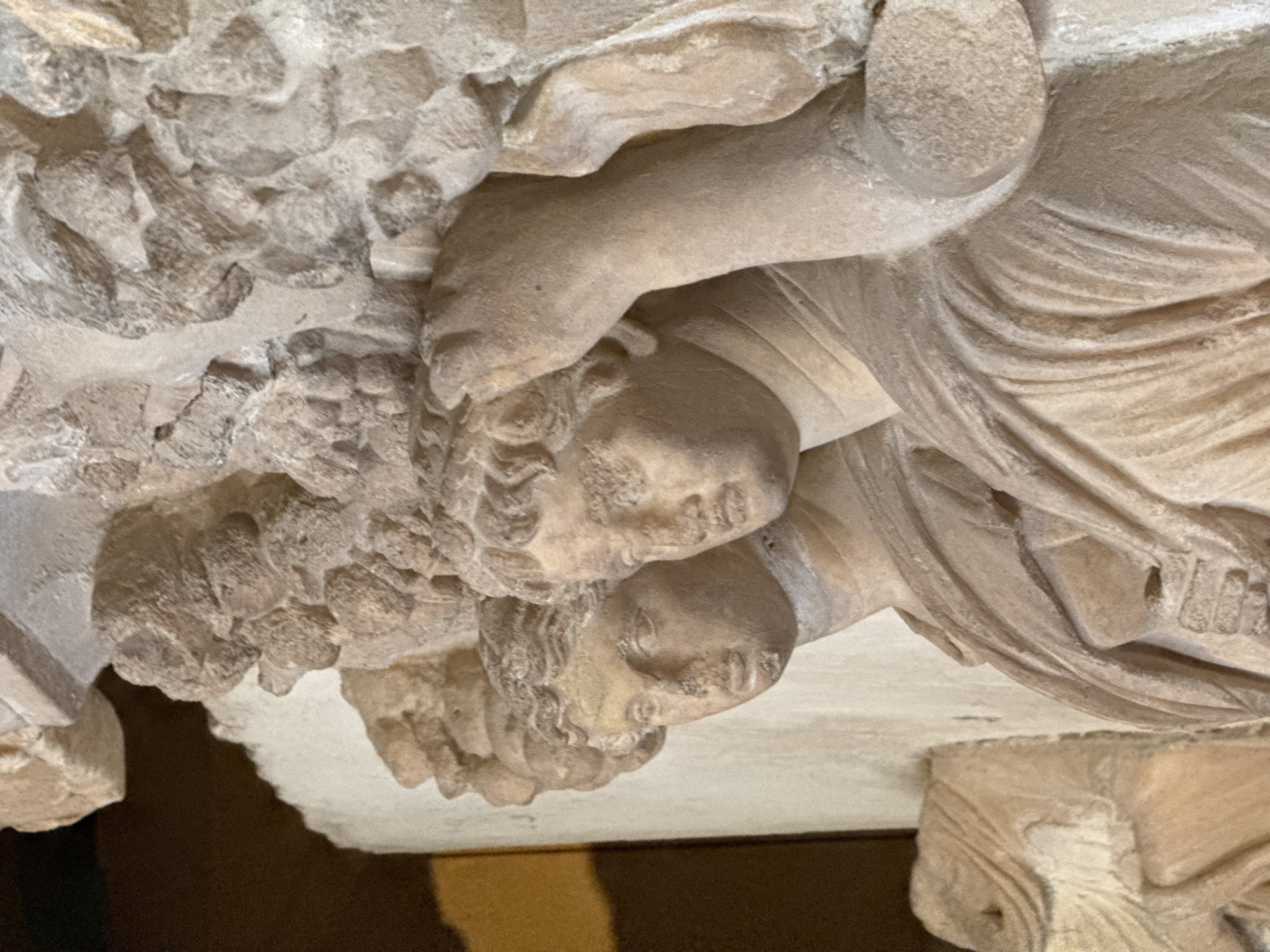 The image depicts a close-up section of an ancient stone carving, possibly a frieze or architectural element. It shows at least two human faces with detailed features and curly hair, surrounded by what appears to be a cluster of leaves or other organic forms. The style suggests a classical or Hellenistic origin.
The carving is located in Pamukkale, Turkey, presumably at the ancient site of Hierapolis, known for its well-preserved ruins including the theater, baths, and necropolis. The specific location within the site is not discernable from the image.