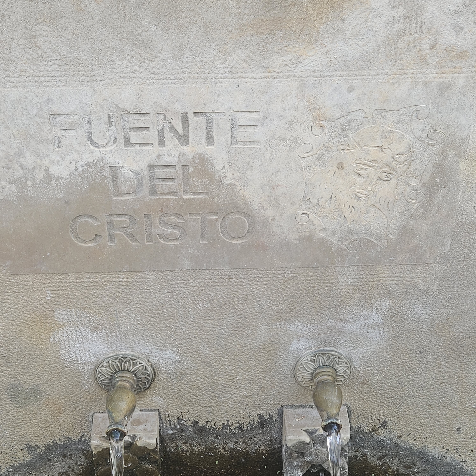 This image depicts a stone wall fountain in Algora, Spain. The inscription "Fuente del Cristo" (Fountain of Christ) is carved into the stone above a stylized image of a man's face. Below the inscription are two metal spouts, each with water flowing from them. The stone around the base of the spouts is stained and darkened.