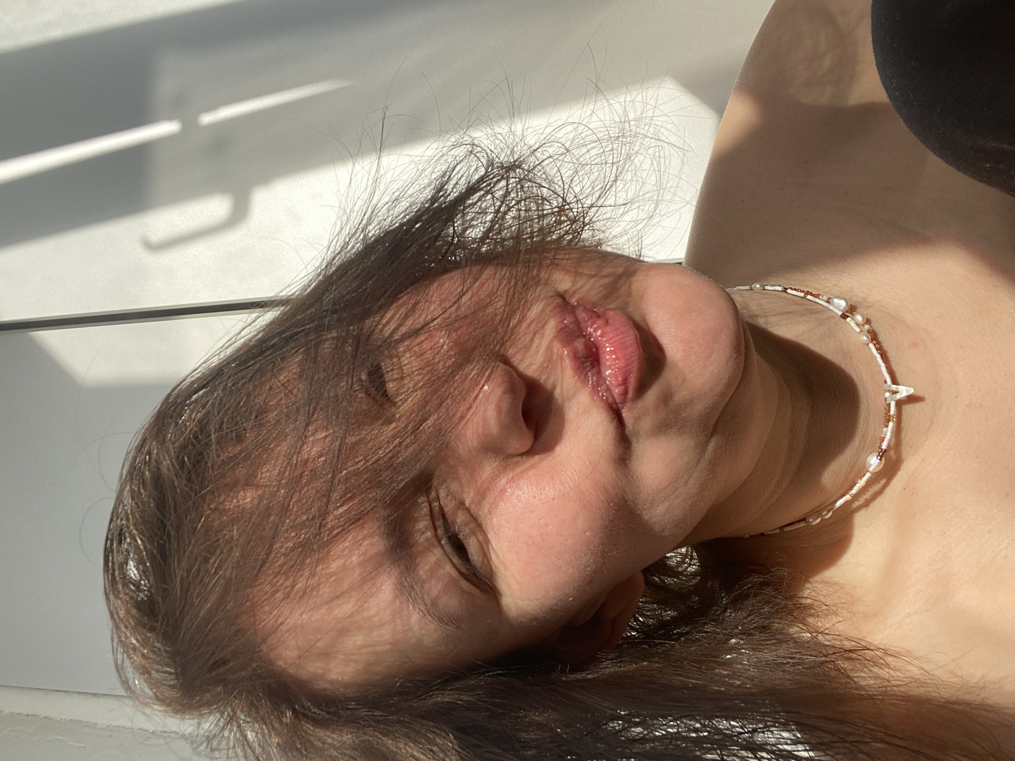 A close-up, high-angle shot captures a person lying down indoors, illuminated by strong natural sunlight. The image is oriented with the person's head tilted to the right, showing their face, neck, and part of their shoulder. The subject has dark brown, slightly disheveled hair that catches the light, creating bright highlights. Their eyes are partially closed or looking downwards, and their lips are slightly parted, revealing a glossy, reddish-pink hue.
A delicate, multi-strand beaded necklace, featuring small white and brown beads along with tiny white pearls, is worn around their neck. Part of a dark-colored garment is visible in the upper right corner, likely a top or strap. The background consists of a bright, light-colored surface, possibly a wall or bed. Strong, directional sunlight casts prominent shadows onto this surface, including distinct horizontal lines suggesting window blinds and a shadow resembling a hook or handle. The harsh lighting creates high contrast, with bright highlights on the person's skin and hair, and deep shadows. The overall impression is one of quiet contemplation or relaxation. No text is visible in the image. The indoor setting lacks specific visual cues for the provided location context of Chaikovsky, Russia.