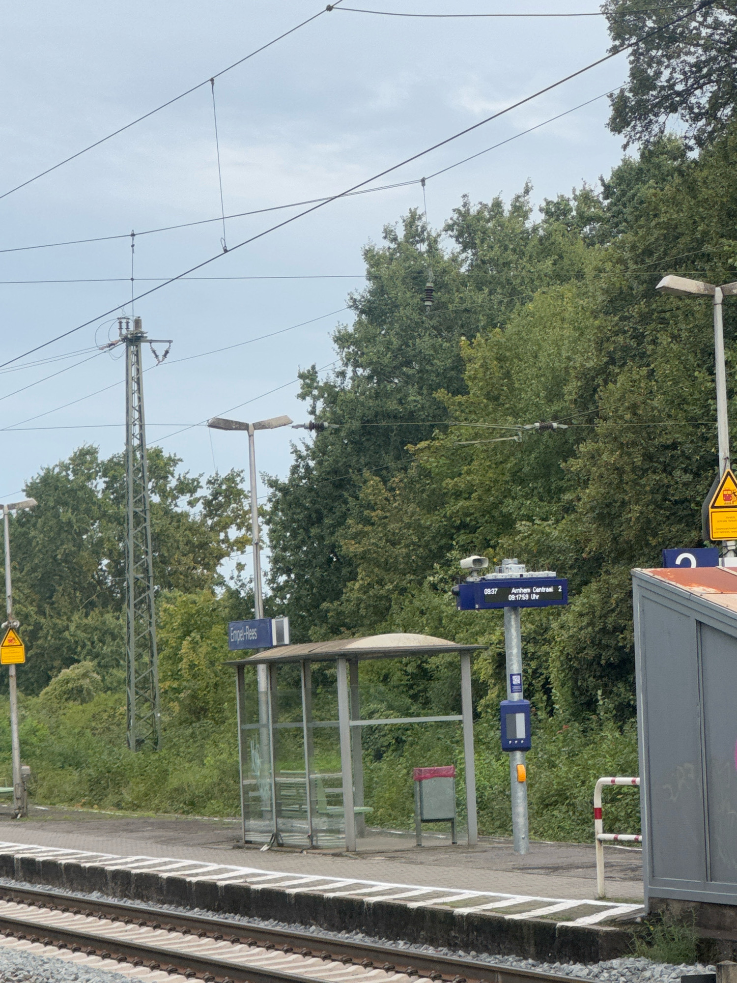 The image shows a train station platform in Rees, Germany. The platform has a small shelter with glass walls and benches inside. There is a blue sign that reads "Empel-Rees". A digital display shows train information, including departure times and destinations, with one entry for "Arnhem Centraal" at "09:37" and another at "09:17:59 Uhr". A tall metal pylon with overhead wires is visible, along with streetlights and vegetation. The train tracks run in the foreground. The sky is overcast. No people are visible in the image.