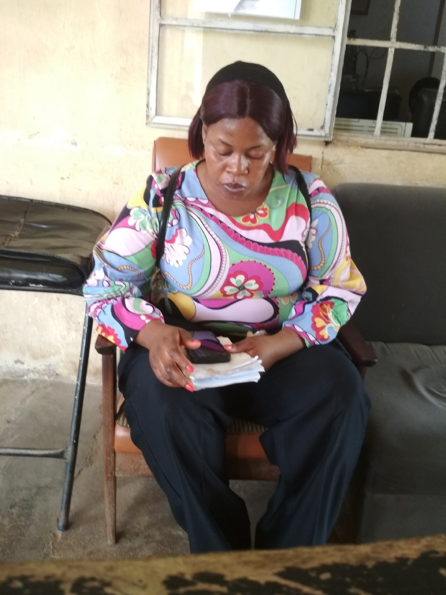 An African woman sits on a wooden chair in what appears to be an indoor waiting area. She is looking down at a stack of papers in her hands, which she is holding with her right hand. The woman has dark skin and dark hair that appears to be styled into a bob with bangs. She is wearing a colorful long-sleeved shirt with a psychedelic pattern, black pants, and her fingernails are painted red. A dark bag is slung over her left shoulder.
The background shows a pale yellow wall with a window that has metal bars. To the left of the woman is a black leather chair or bench, and to her right is a dark gray upholstered sofa. The floor is concrete. The scene suggests a government office, clinic, or other public building in Bisichi, Nigeria.