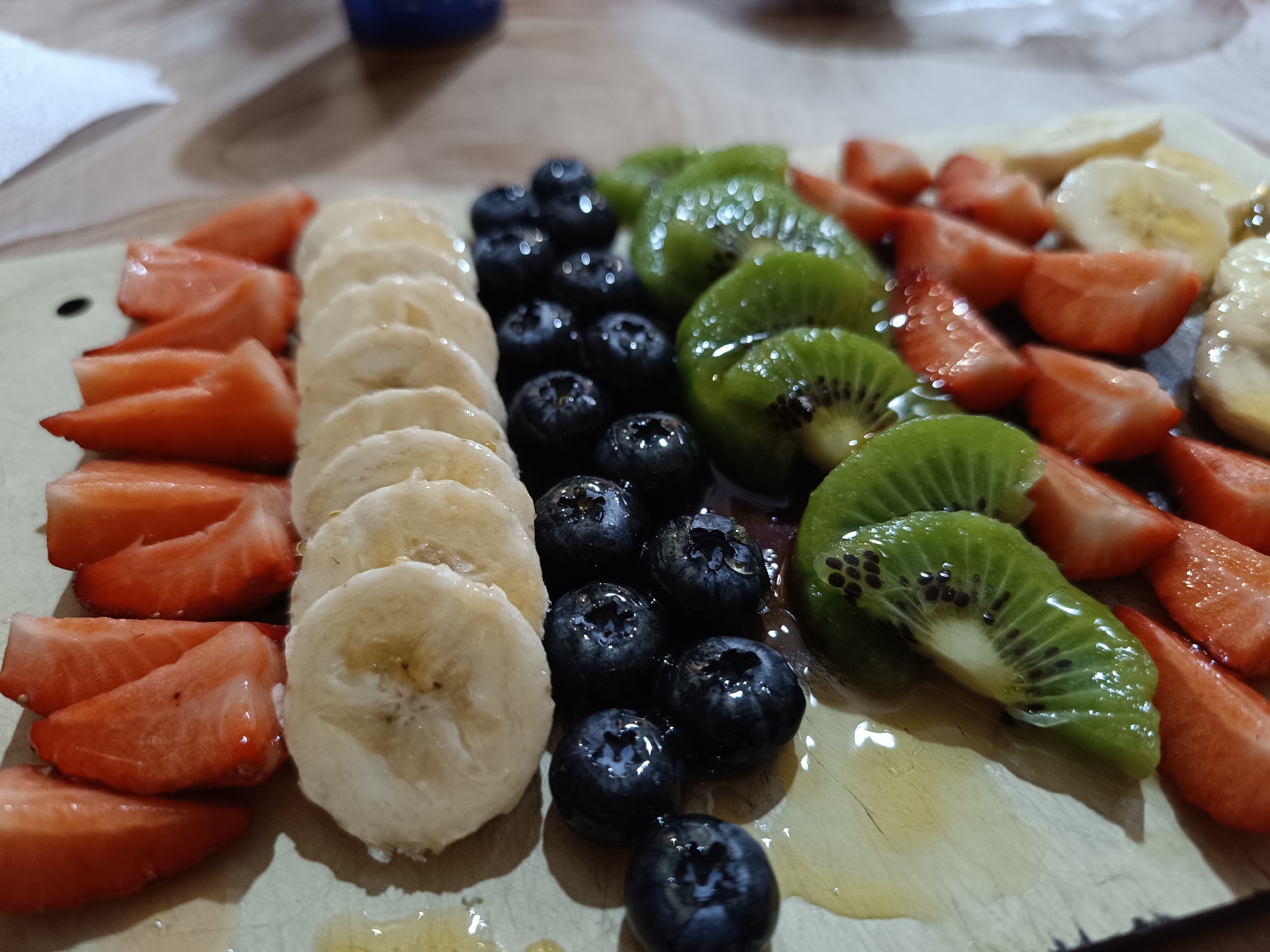 The image shows a close-up of a wooden platter arranged with various fruits. The fruits are arranged in rows, including sliced strawberries, banana slices, blueberries, and kiwi slices. There appears to be a clear glaze or syrup drizzled over the fruit. The location and city of Pleven, Bulgaria cannot be determined from the image content.