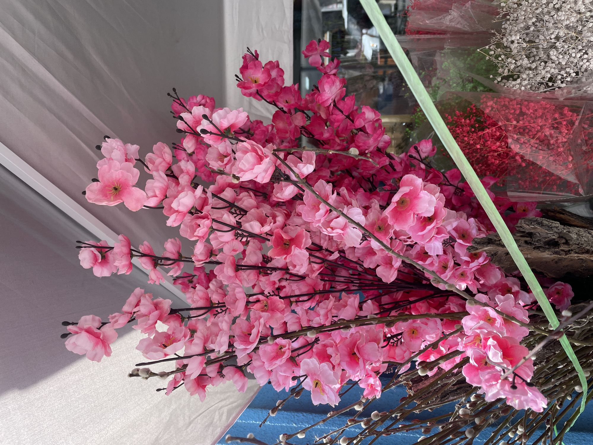 The image shows a display of artificial pink cherry blossom branches. The flowers are clustered densely on dark brown stems. At the bottom are pussy willow branches with greyish-white catkins. In the background, through a window, are other bouquets of dried flowers, including red and white baby's breath. There are green floral stems, and a piece of tree bark can be seen. The photo appears to be taken indoors, possibly in a flower shop, with light shining on the white fabric. It is unknown if this is in Yerevan, Armenia.