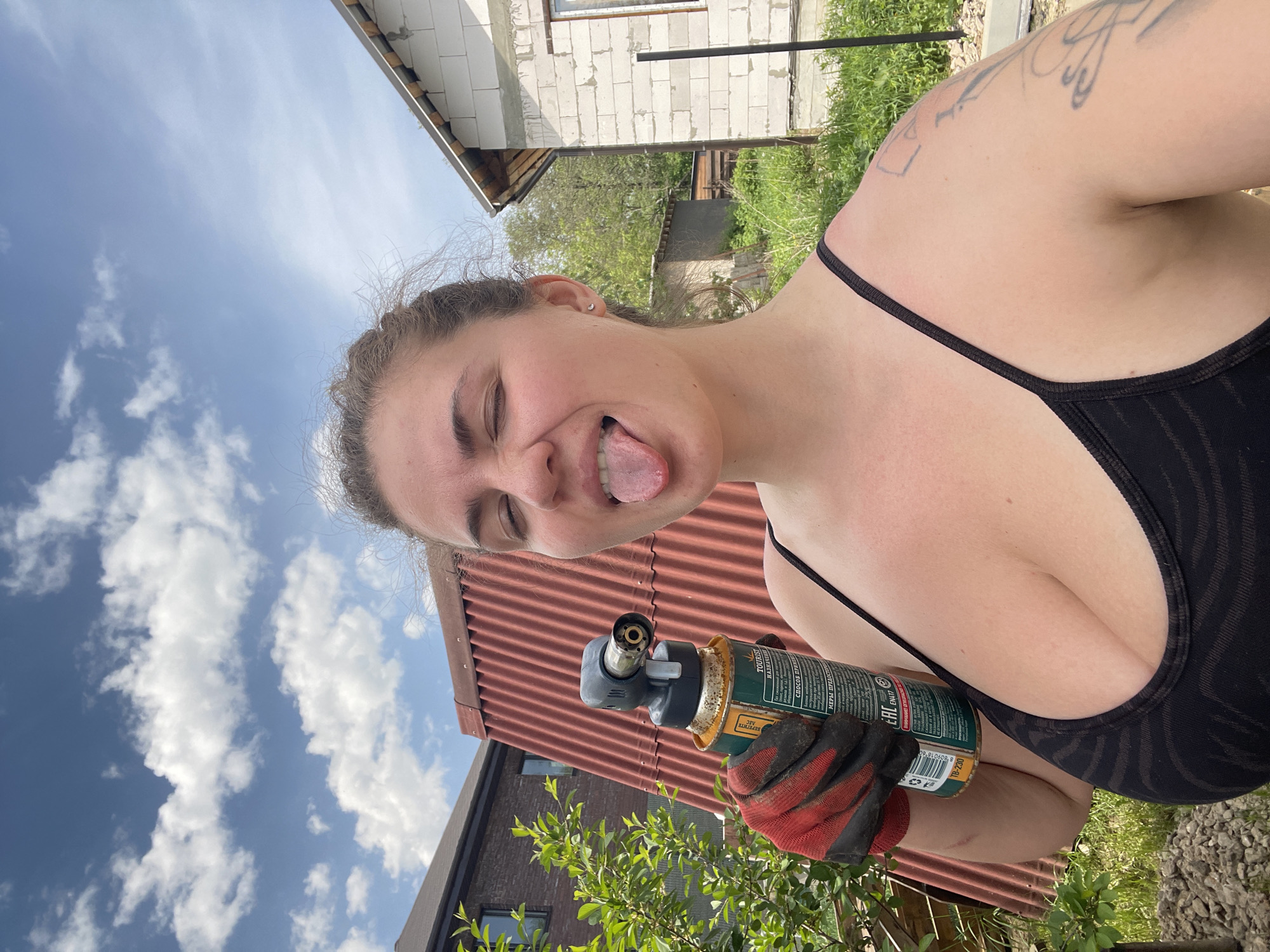 A woman is holding a propane torch. She is sticking her tongue out and winking. She is wearing a black tank top and red and black work gloves. The location is outdoors, with a red metal roof and a building made of white blocks visible in the background.