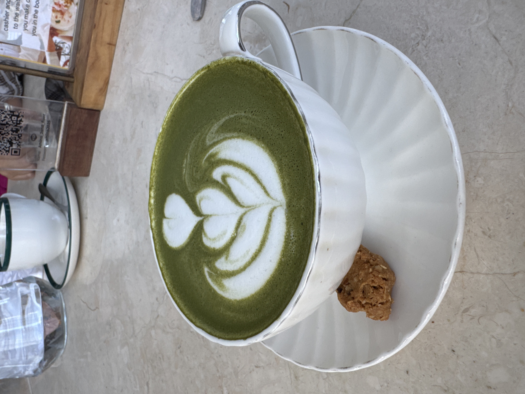 A high-angle shot shows a cup of matcha latte with latte art on a white saucer, accompanied by a small cookie. The cup and saucer are placed on a marble-like surface next to a small table with a menu stand and another cup in the background. The scene appears to be set in a café or restaurant, and the photo was potentially taken in Ungasan, Indonesia, based on the user prompt.