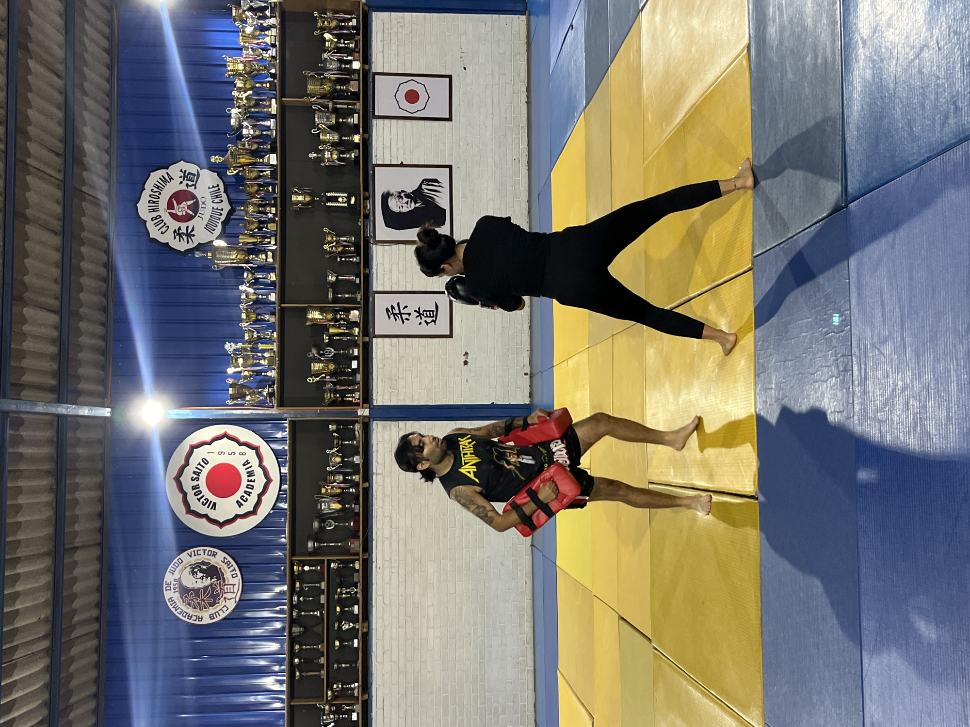 The scene depicts a martial arts training session within a dojo identified as Club Hiroshima Judo and Victor Saito Academia in Iquique, Chile. The room features blue corrugated metal walls on the left and a white brick wall on the right, with a corrugated metal ceiling. The floor is covered with interlocking blue and yellow foam mats. Bright overhead lights cast distinct shadows.
The background walls are heavily decorated. On the left, two large circular signs are visible: one for "Academia de Judo Club Victor Saito 1958" featuring a portrait, and another for "Victor Saito Academia 1958" with a red sun symbol. A central white sign reads "CLUB HIROSHIMA JUDO IQUIQUE CHILE" with a traditional judo symbol. Numerous gold and silver trophies line shelves beneath these signs, extending across the entire back wall. On the white brick wall to the right, three framed items are displayed: Japanese characters "柔道", a black and white portrait of an elderly man, and a white flag with a red circle.
Two individuals, an adult male and an adult female, are positioned in the center of the mats, both barefoot. The male, with dark hair tied back and visible arm tattoos, wears a black sleeveless shirt with "ANTHRAX" text and black athletic shorts. He holds a red kick pad in each hand, facing the female. The female, with dark hair pulled into a bun, wears an all-black long-sleeved shirt and black athletic pants. She wears black boxing gloves and stands in an aggressive fighting stance, facing the male, with her gloved hands raised. The interaction indicates a pad work training exercise.