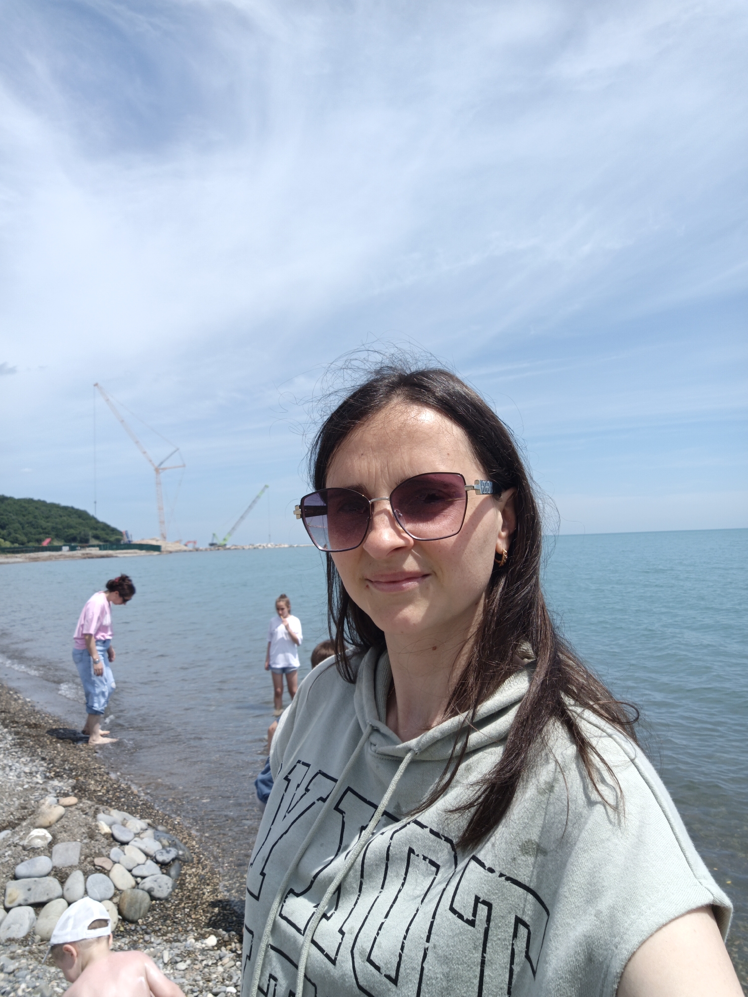 A female, likely in her 30s or 40s, with long dark hair and reflective sunglasses, is positioned in the foreground, facing and smiling slightly towards the camera. She wears a light green or grey sleeveless hooded sweatshirt with dark lettering. Her left ear displays a small golden hoop earring. The scene is a pebbly beach under a clear blue sky with sparse white cirrus clouds, indicative of sunny weather. Calm, blue-green water extends from the beach into the distance. In the left mid-ground, a woman with dark hair in a pink t-shirt and light blue jeans stands barefoot in the shallow water, looking downwards. Further right, a young girl in a white t-shirt and shorts stands in the water. In the bottom-left, a young child wearing a white baseball cap is partially visible on the pebbly shore, facing away from the camera. On the distant horizon, a green, tree-covered hill is present on the left, and multiple large construction cranes, including a tall yellow/orange lattice boom crane and a smaller green boom crane, are visible over the water, suggesting ongoing construction or port activity. A low barrier or breakwater extends from the land into the water near the cranes. The location is Krinitsa, Russia.