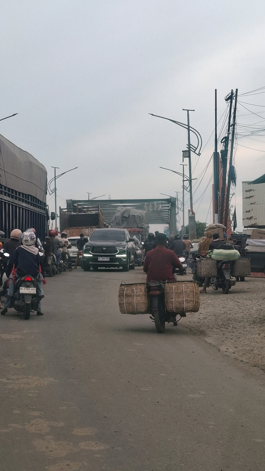 The image depicts a street scene in Meuse, Indonesia, characterized by moderate traffic congestion. In the foreground, a motorcycle with a rider wearing a red shirt is moving away from the camera, carrying two large woven baskets on its rear. To the left, another motorcycle with a rider in a blue shirt and a helmet is also visible. The middle ground features a dark-colored SUV with its license plate visible as "BL 1900 AZD", also amidst traffic. Several other motorcycles and vehicles are present, suggesting a busy road. In the background, trucks and what appear to be stalls or buildings line the street. The sky is overcast, indicating a cloudy day. The road surface appears to be asphalt, with some patches and debris visible. The overall atmosphere is that of a typical urban environment with daily transportation activities.