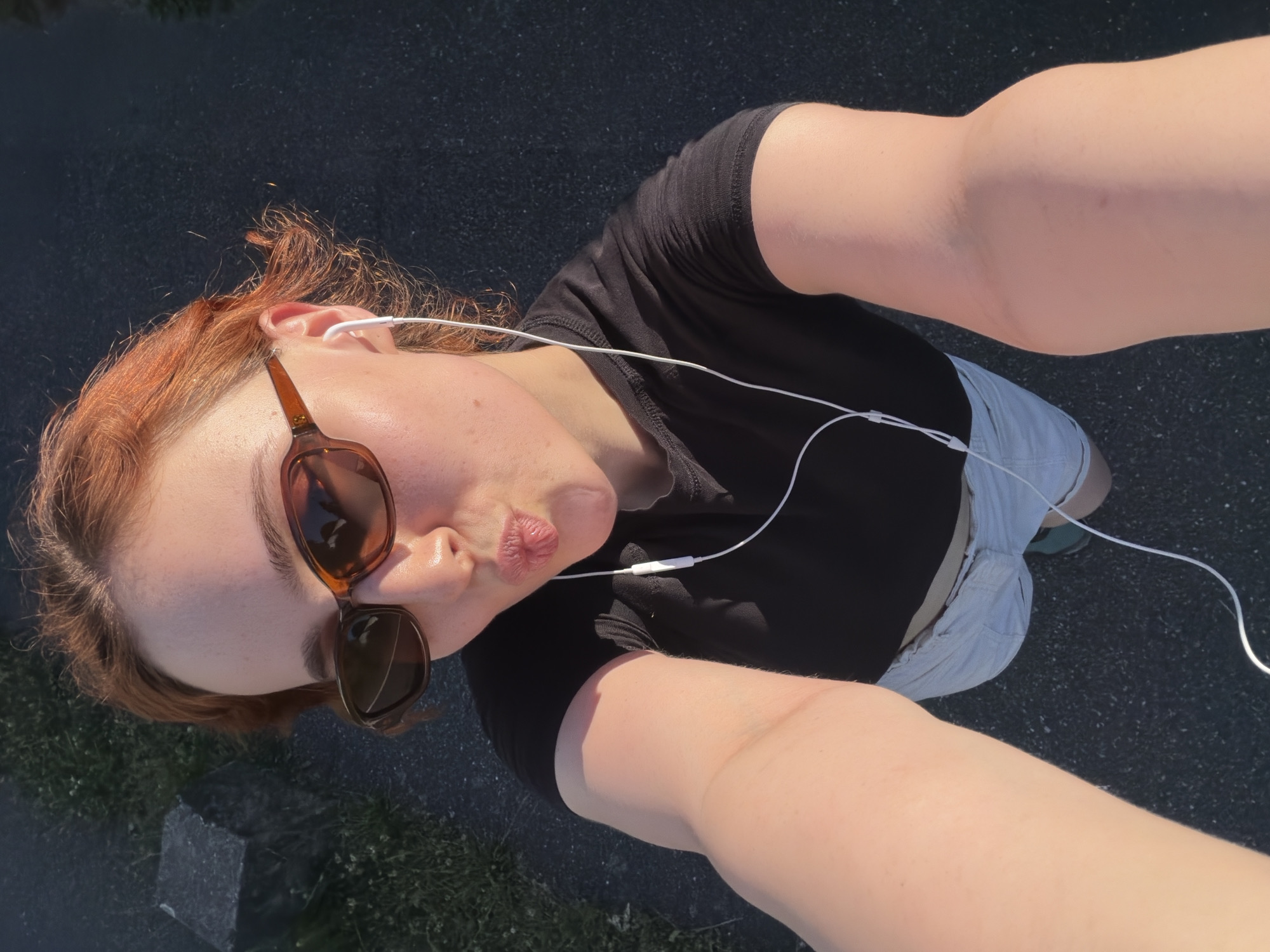 A person with auburn hair is taking a selfie outdoors. They are wearing sunglasses, a black t-shirt, and light-colored shorts. White earbuds are in their ears. Their lips are puckered. The ground is dark asphalt, with some patches of grass and a large dark stone visible in the background. The lighting suggests it is late afternoon or early evening. The scene does not contain any identifiable landmarks or elements that would indicate a specific location within the city of Strasbourg, France.