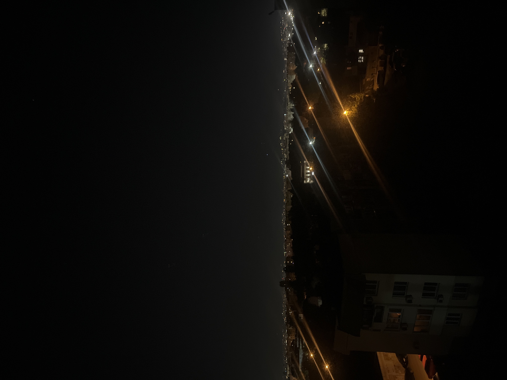 A nighttime shot shows a dimly lit cityscape next to a body of water. The right side of the image shows buildings, with a large white building in the foreground featuring multiple lit windows. Streetlights cast long, angled beams across the scene. The left side presents a dark, open expanse, presumably a body of water with very few visible lights. The photo appears to be taken from an elevated position, potentially a high-rise building or vantage point, overlooking the city and the water. The location is unknown, but is NOT in Sofia, Bulgaria.