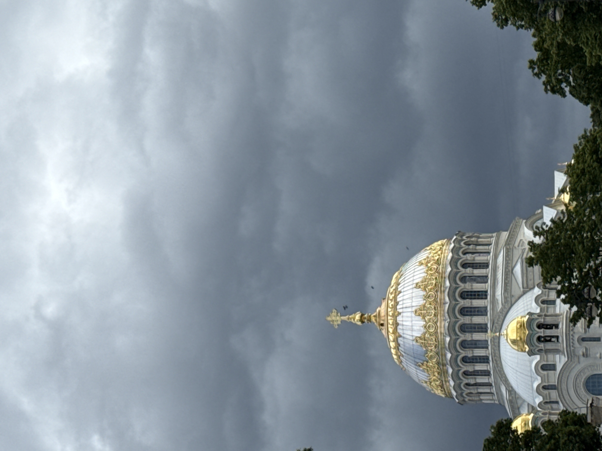 The image shows a close-up, low-angle view of the Naval Cathedral of Saint Nicholas in Kronstadt, Russia, under a cloudy sky. The cathedral's prominent dome, adorned with gold ornamentation and a cross, is the central focus. The building's white facade and intricate architectural details are visible, along with smaller golden domes and the surrounding green trees. The sky is filled with heavy, dark gray clouds, suggesting an overcast or potentially stormy day. There are no people visible in the frame, and no discernible actions or interactions are taking place. The scene is static and focuses solely on the architecture of the cathedral and the atmospheric conditions.