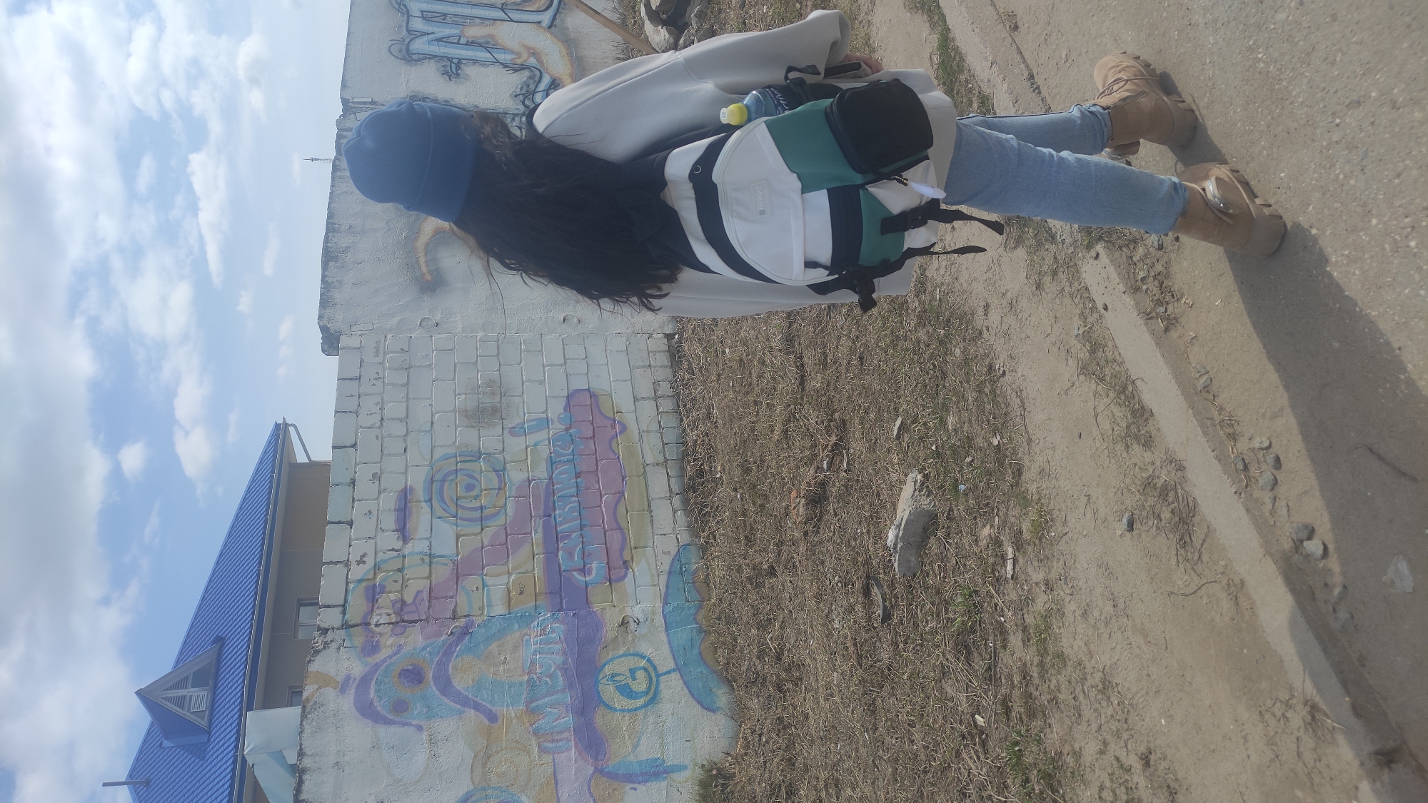 The image, rotated 90 degrees counter-clockwise, captures a person from behind, standing outdoors in what appears to be Chaikovsky, Russia, on a bright day. The individual has long dark hair and is wearing a blue beanie-style cap, a light-colored (possibly white or cream) long-sleeved top or jacket, light blue jeans, and tan/brown work-style boots. They are carrying a two-tone backpack, predominantly white with dark green sections and black straps, with a water bottle visible in a side pocket.
The person is standing on a dirt path with sparse dry grass and small rocks, alongside a concrete curb. To their left is a white brick or block wall, weathered and adorned with colorful graffiti featuring abstract patterns in shades of light blue, purple, and yellow. In the background, a building with a distinctive blue-tiled roof and an attic window is visible, set against a bright sky with scattered white clouds. The scene suggests clear daytime conditions. There is no specific activity depicted other than the person standing or looking towards the left, and no visible text is clearly legible on any object or surface.