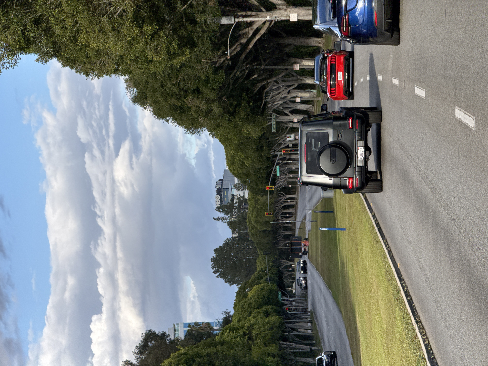 The image shows a north-facing view of Hillcrest Road in Beverly Hills, California. A line of vehicles is stopped on the road, including a black Mercedes-Benz G-Wagon, a red Ford Mustang, and a blue BMW. The road is lined with trees that form a canopy overhead. There are traffic lights visible along the road, all showing red. A green grassy median separates the lanes of traffic. A street sign for "Hillcrest Rd" is also visible. In the distance are some buildings and a cloudy sky.