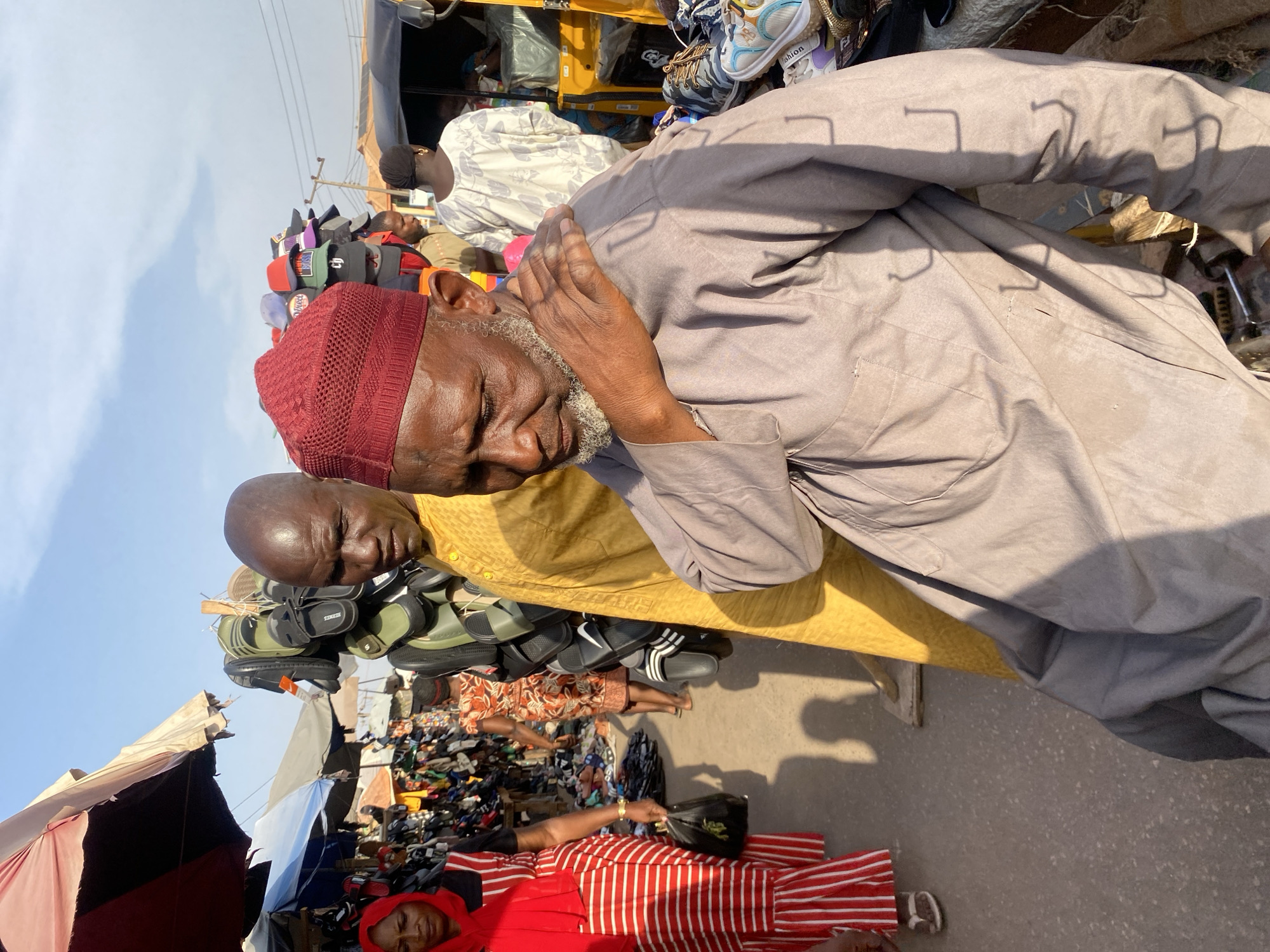 The scene depicts an outdoor market in Sabon Tasha, Nigeria, under bright daytime lighting conditions. In the immediate foreground, an elderly man with a white beard and a maroon knitted kufi cap wears a light brown long-sleeved garment, his right hand resting on his left shoulder, with his gaze directed downwards. To his left and slightly behind, a bald man in a yellow patterned kaftan stands, looking forward, positioned near a vertical display of olive green and black sandals hanging from hooks. Further left, a woman wearing a red hijab and a red and white striped dress walks away from the viewer, holding a black plastic bag.
The market environment is densely packed with merchandise and people. Various shoes, slippers, and sneakers are displayed on stands, racks, and laid out on the ground. A prominent display of colorful baseball caps is visible in the background behind the elderly man. Overhead, large canvas umbrellas and awnings, including red, blue, and grey ones, provide shade. A yellow motorized tricycle (auto-rickshaw) is partially visible on the far right. Several other individuals are present in the midground and background, moving through the market. The ground is paved. Overhead, power lines and a utility pole are visible against a light blue sky with scattered clouds. Strong sunlight creates distinct shadows, including hook-shaped shadows on the elderly man's garment.